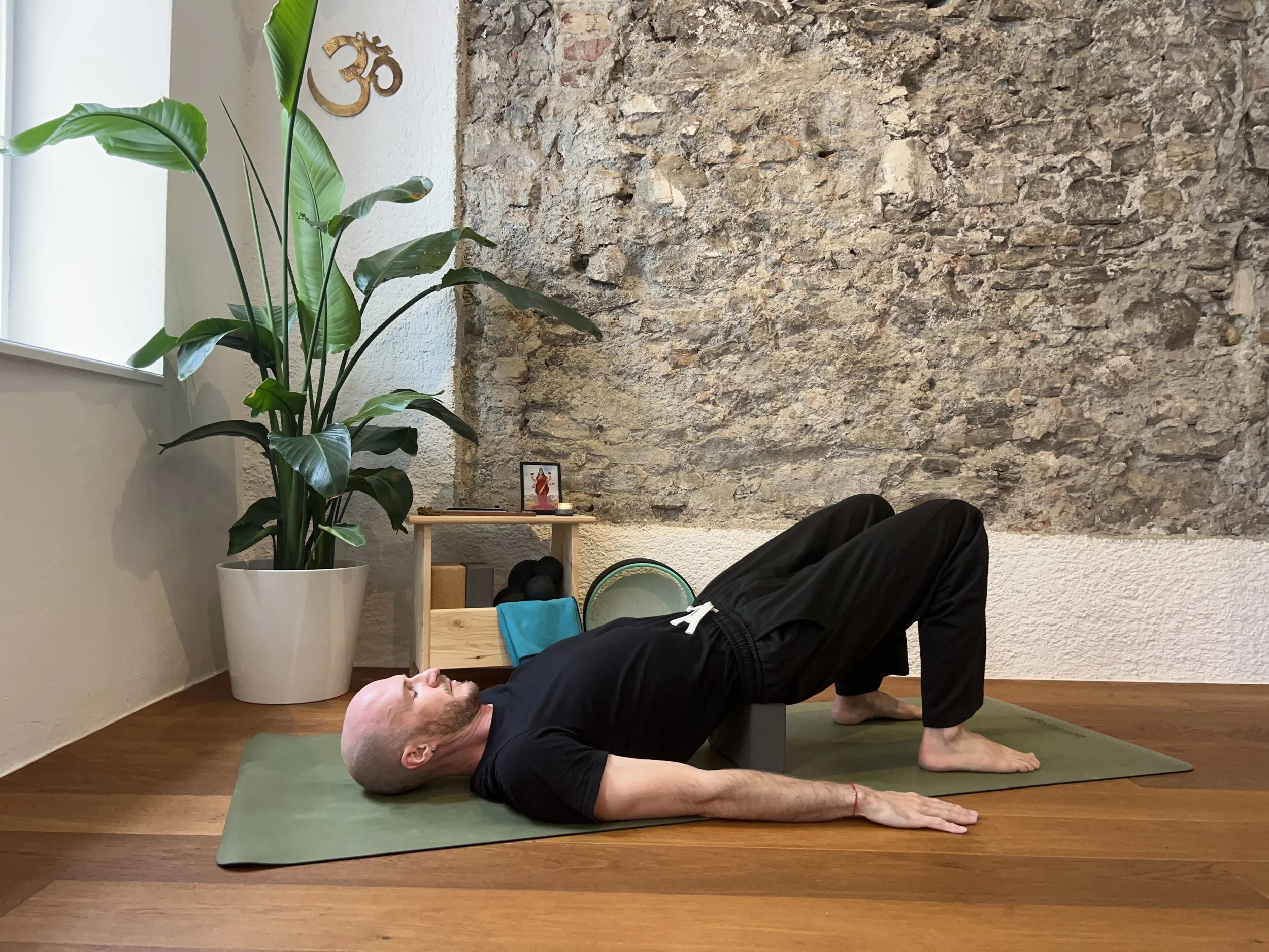 Gregor Eistert practicing bridge pose on a green mat in a room with a large plant in a white pot, a textured stone wall, and a small altar with a picture of a deity, candles, and a small altar table.