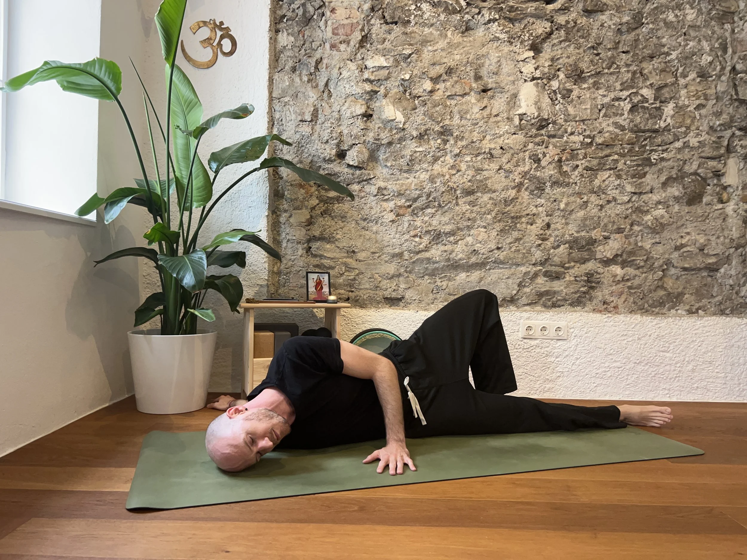 Gregor Eistert practicing broken wing pose in a room with a large brick wall, a potted plant, and a small altar with a picture and a candle. He is lying on a green yoga mat, with his head resting on his hands, and his eyes closed.