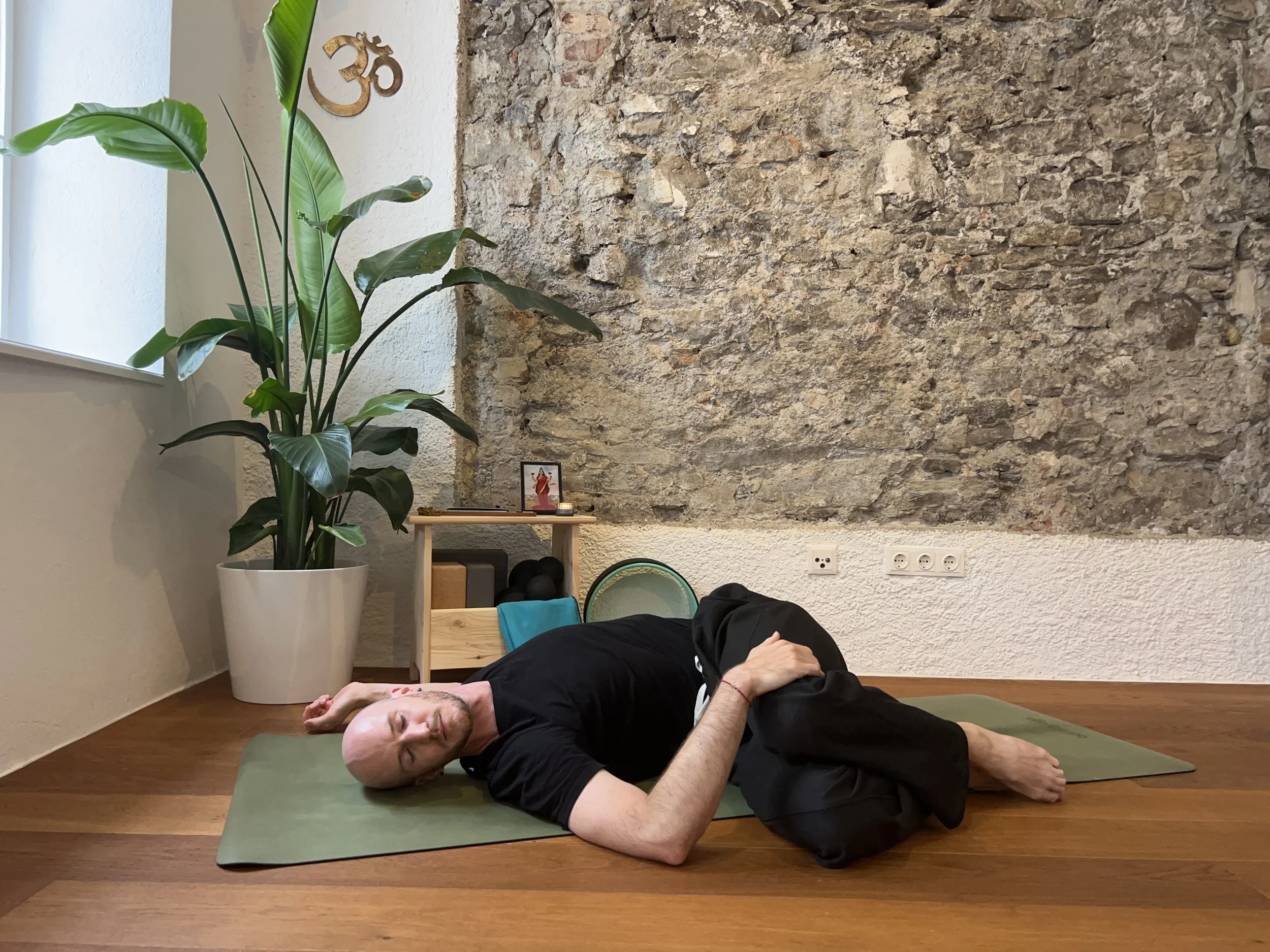 A man with a shaved head lying on his side on a green yoga mat, eyes closed, relaxing in a room with a brick wall and a large green plant in a white pot, with a small altar or shelf with a picture, candles, and functional electrical outlets in the background.