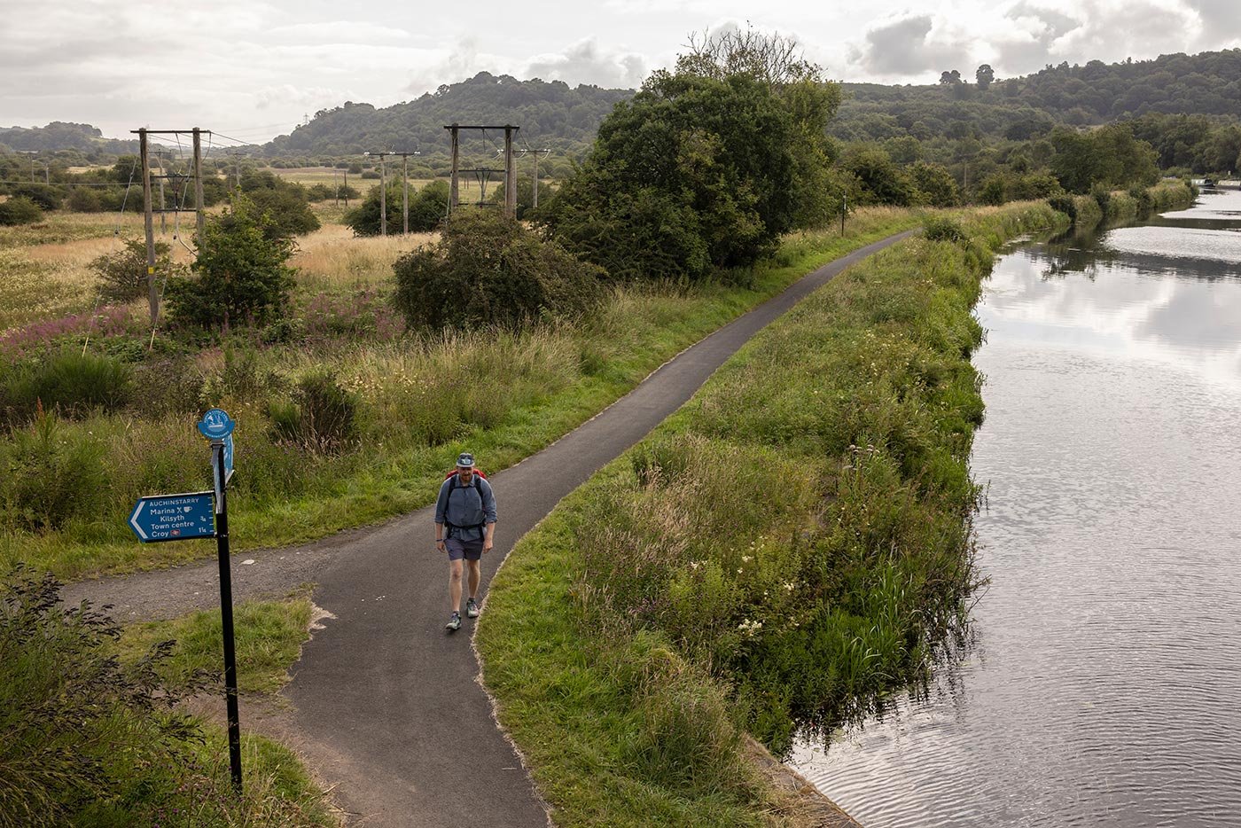 A man walks towards the camera, along a canal path,. A blue sign points ina. few directions, and to the right of image is the canal water.