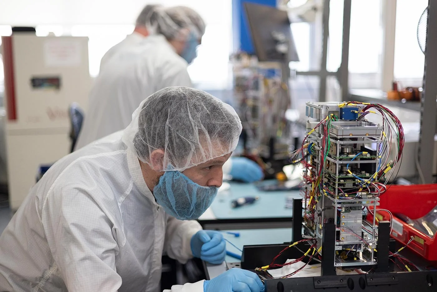 Man wearing lab coat and hair net looks at a stack of technology and wires, a satellitle being built. He's backlit in a science lab type environment.