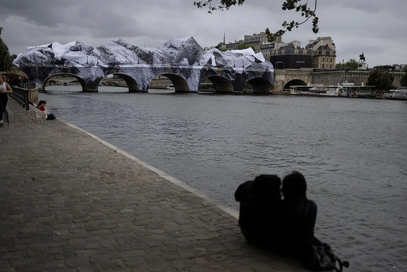 A couple sit in silhouette on a riverbank, beside a grey river. IN the distance behind them is an inflatable artwork which wraps a whole bridge. Buildings are visible on the top right of the image. The wrapped bridge is a striking contrast to scene.