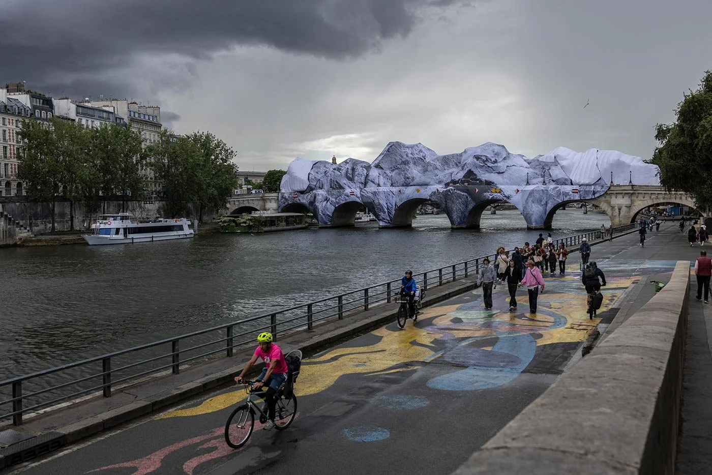 People cycle and walk along a river bank quay, a grey river to the left of image. IN disctance an inflatable artwork in black and white envelopes a bridge.