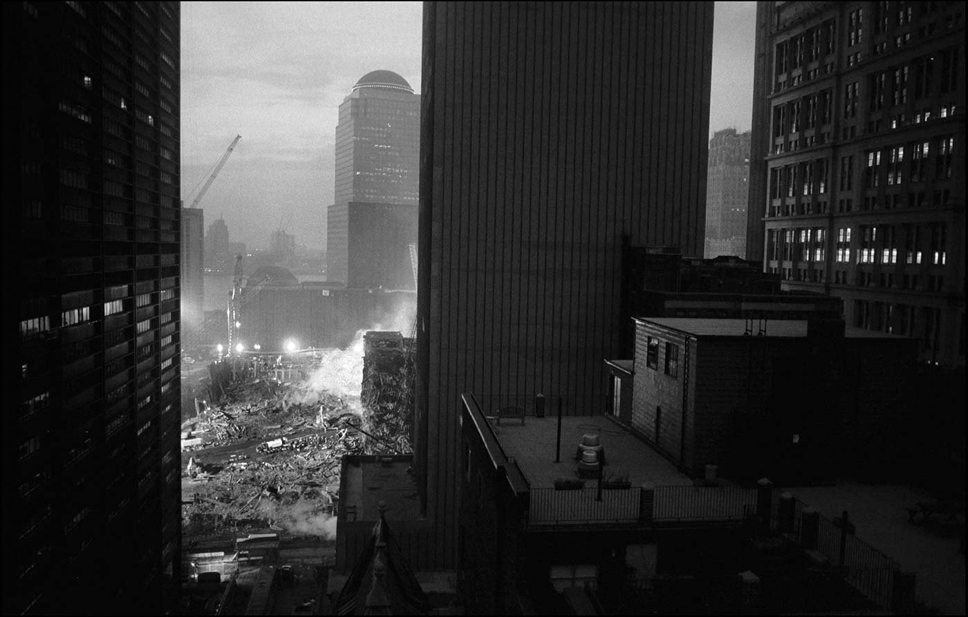 Black and white photo showing the devastation at Ground Zero in Manhattan, at dusk. Nearby buildings have their office lights on, and down below the Ground Zero site.