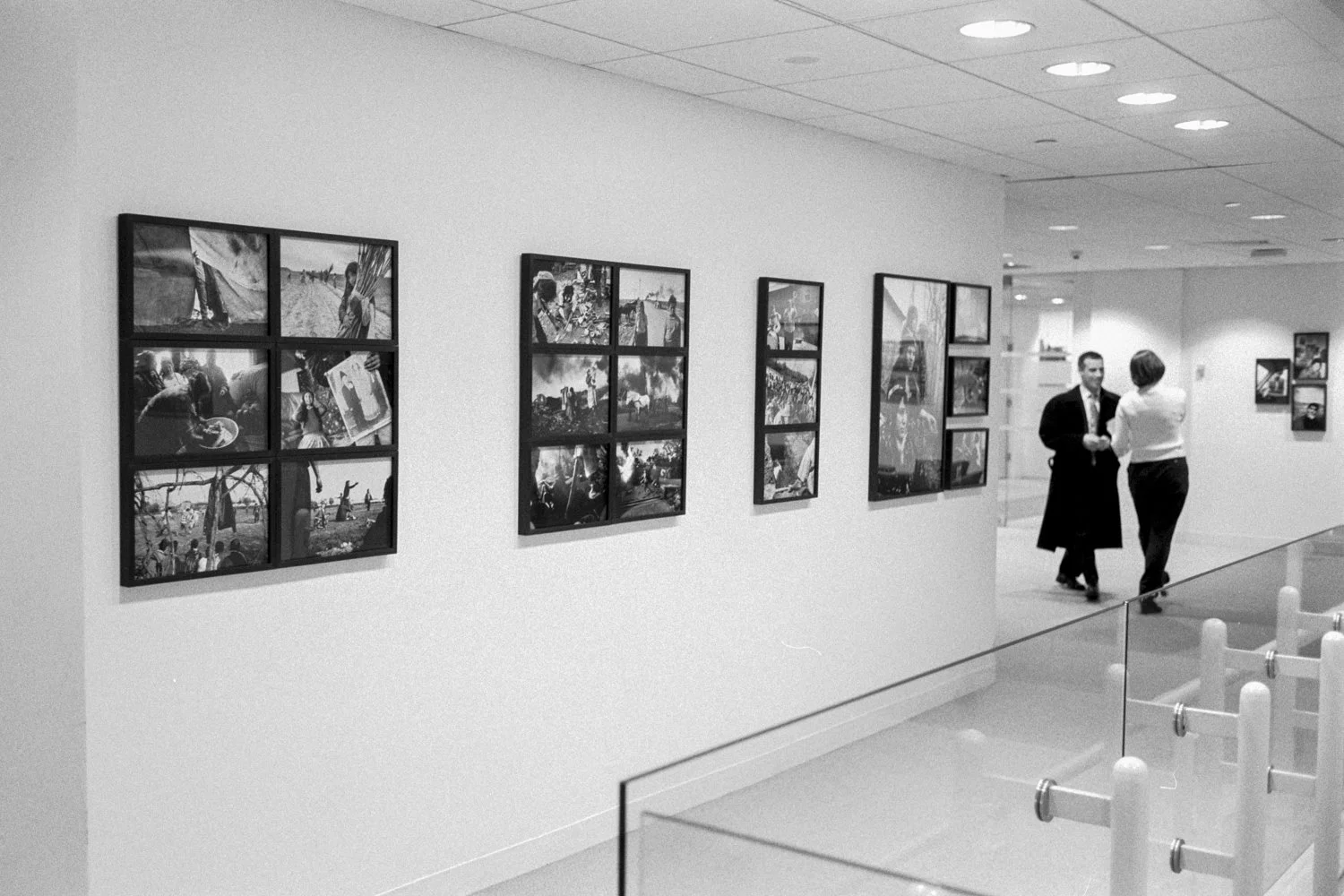 Black and white frames photographs hang on white office walls, as an exhibition, and in background two people slightly out of focus.