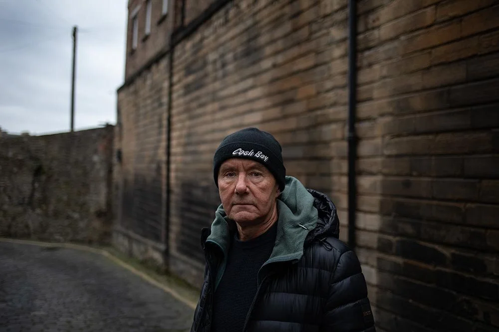 A serious looking man stands infront of a large wall, he wears a had on which it reads Cosh Boy, and he wears a dark puffer jacket.