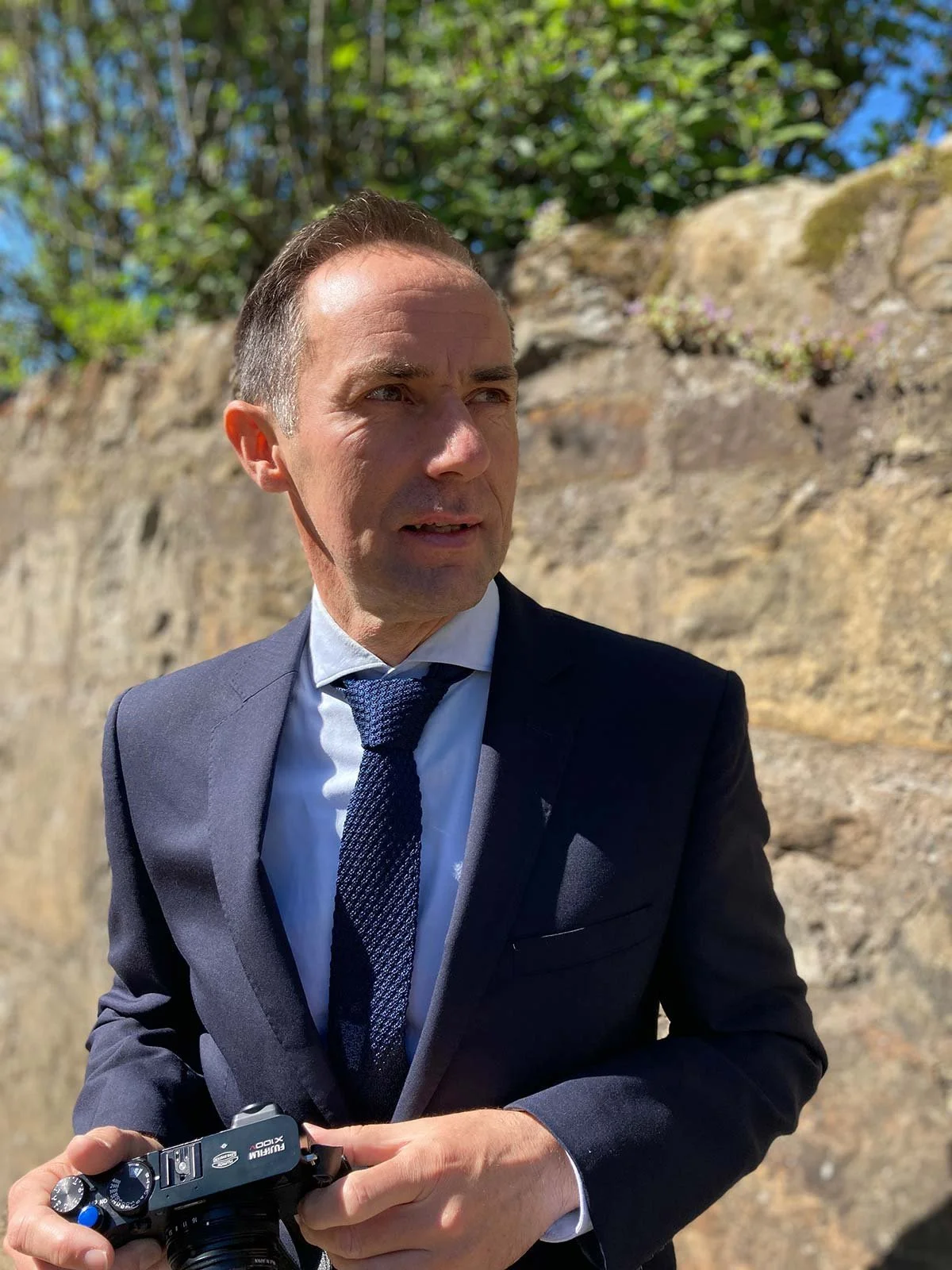 A man in a dark business suit holding a camera, standing outdoors on a sunny day with a stone wall and green foliage in the background.