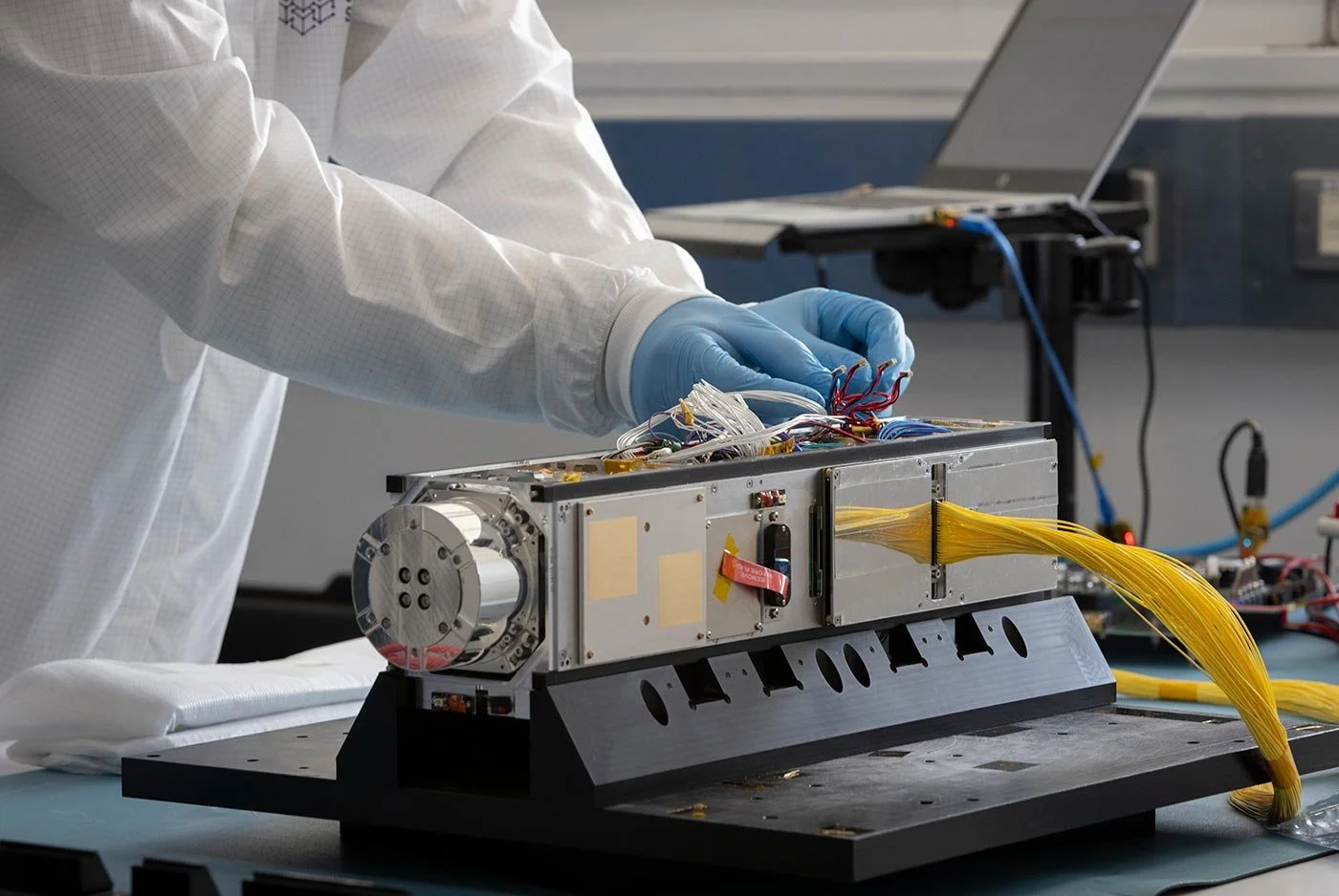 Pair of hands wearing blue gloves, and person wearing white lab coat, toucing wires of a small CubeSat satellitle, in a clean room of a technology centre.