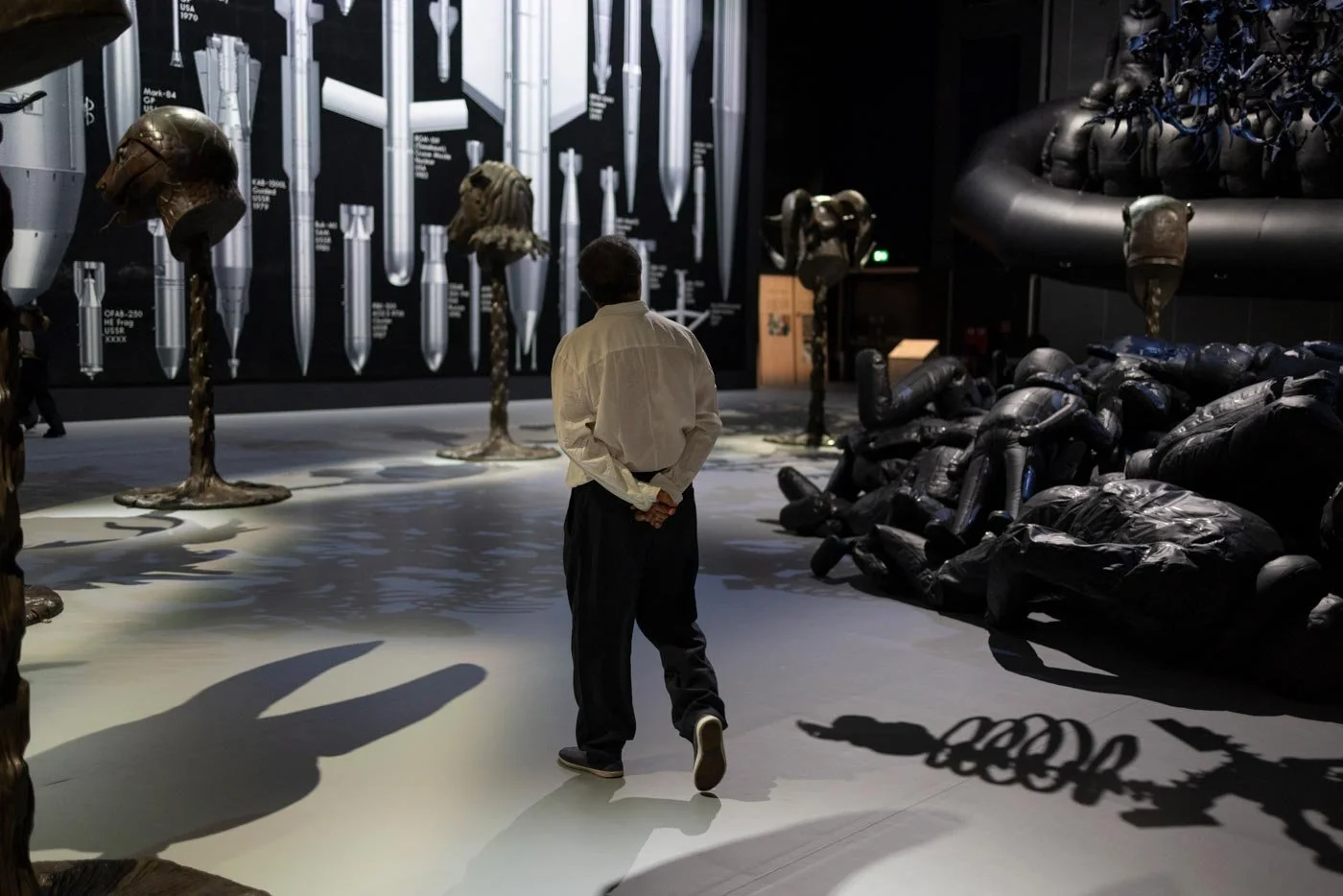 Chinese artist Ai Weiwei wearing a white shirt walks away from the camera, through an exhibition of installations and sculptures.