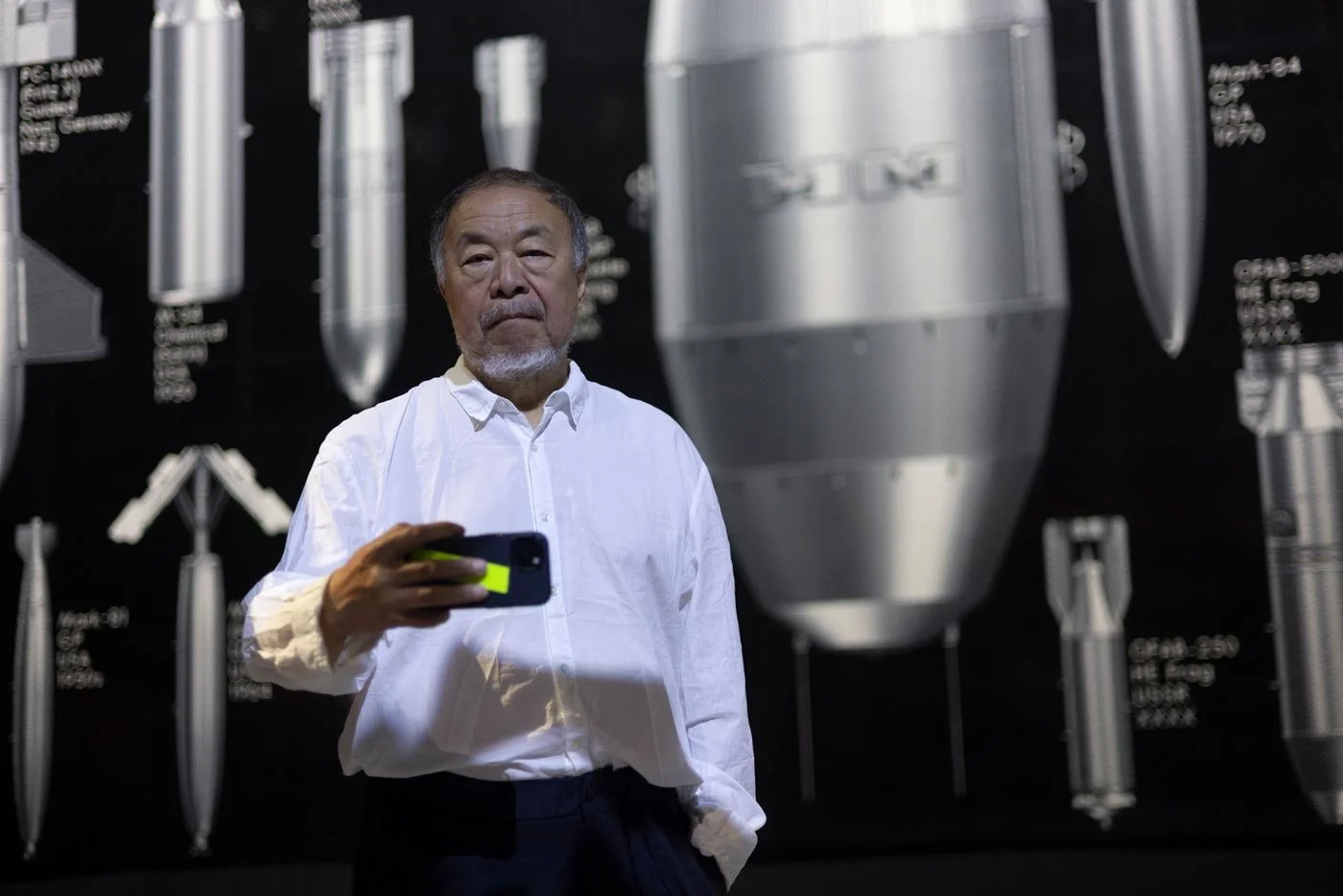 Chinese artist Ai Weiwei wearing a white shirt walks stands in front of an artwork depicting bombs, he uses his smartphone to photograph the photographer.