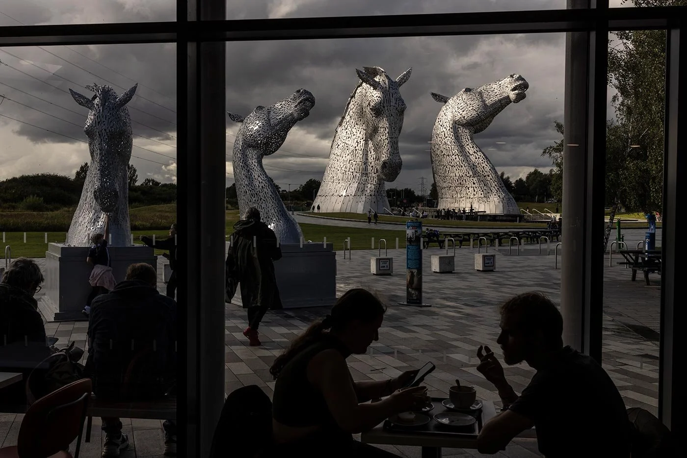A view of the Kelpies monumental horse sculptures, seen from inside the visitor centre cafe. The sculptures are lit very dramatically by light and dark stormy clouds.
