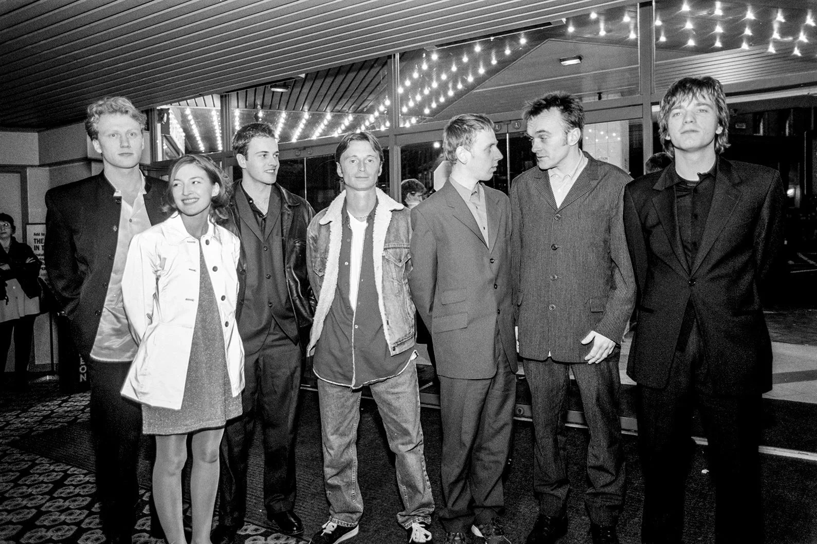 A line up of actors of the movie Trainspotting, standing inside a cinema, looking at the cameras, there are 6 men and one woman. The photo is in black and white.