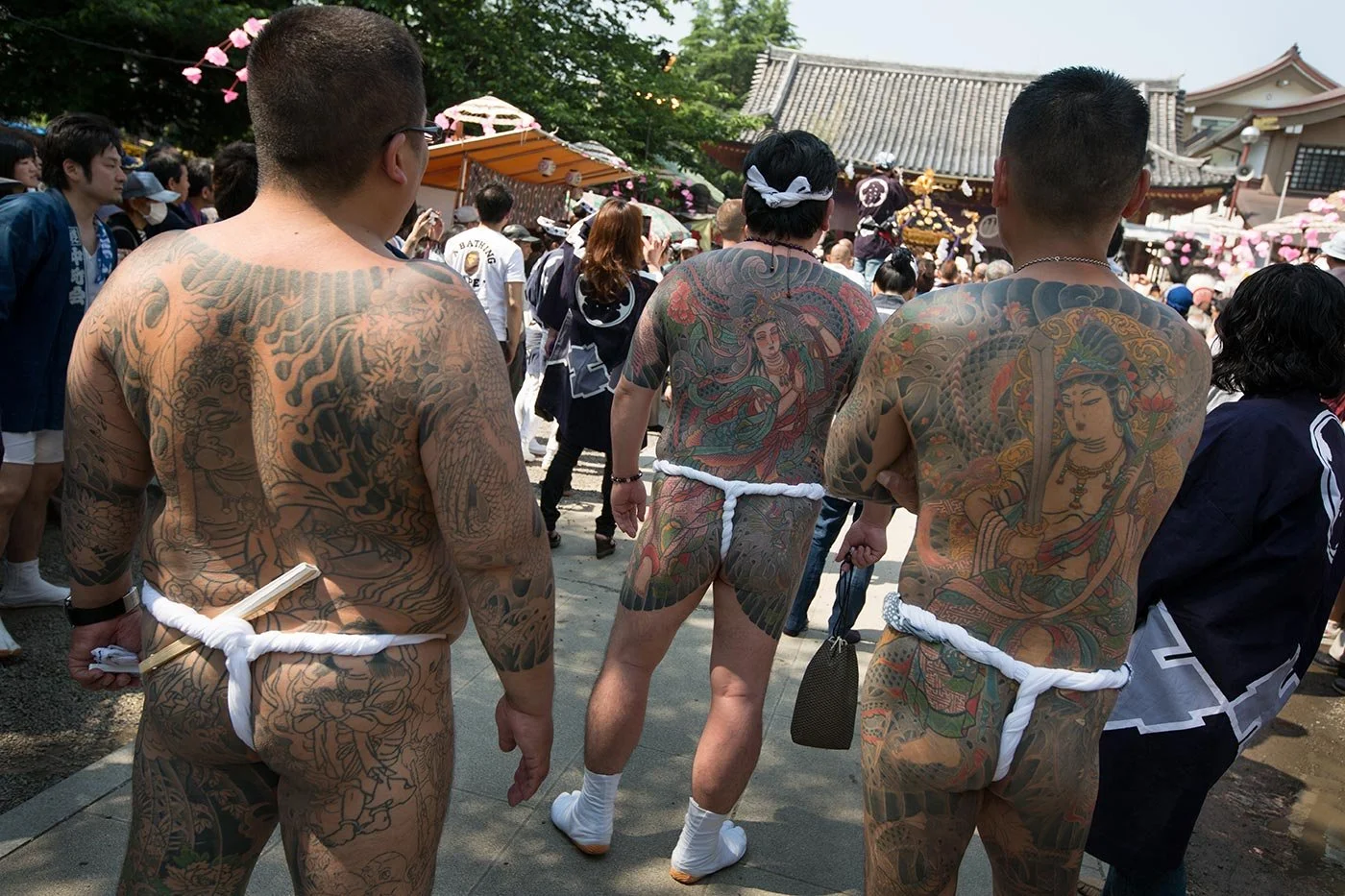 Three men stand naked, with their heavily tattooed backs to the camera