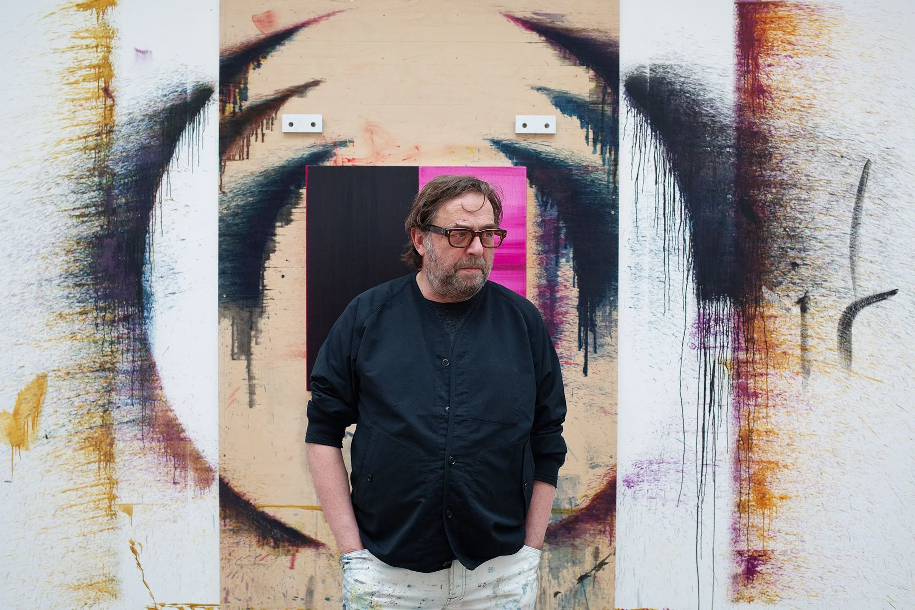 An artists stands in front of a paint splattered wall in his studio. He wears dark shirt and glasses.