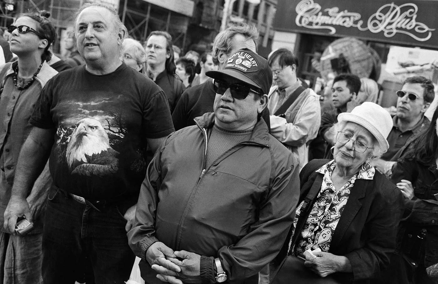 Black and white photo of a group of people, one man wears a tshirt with an American Giolden Eagle on it, another man wears a skip cap and and watch on outside of his jacket wrist, and beside them an old lady in white hat.