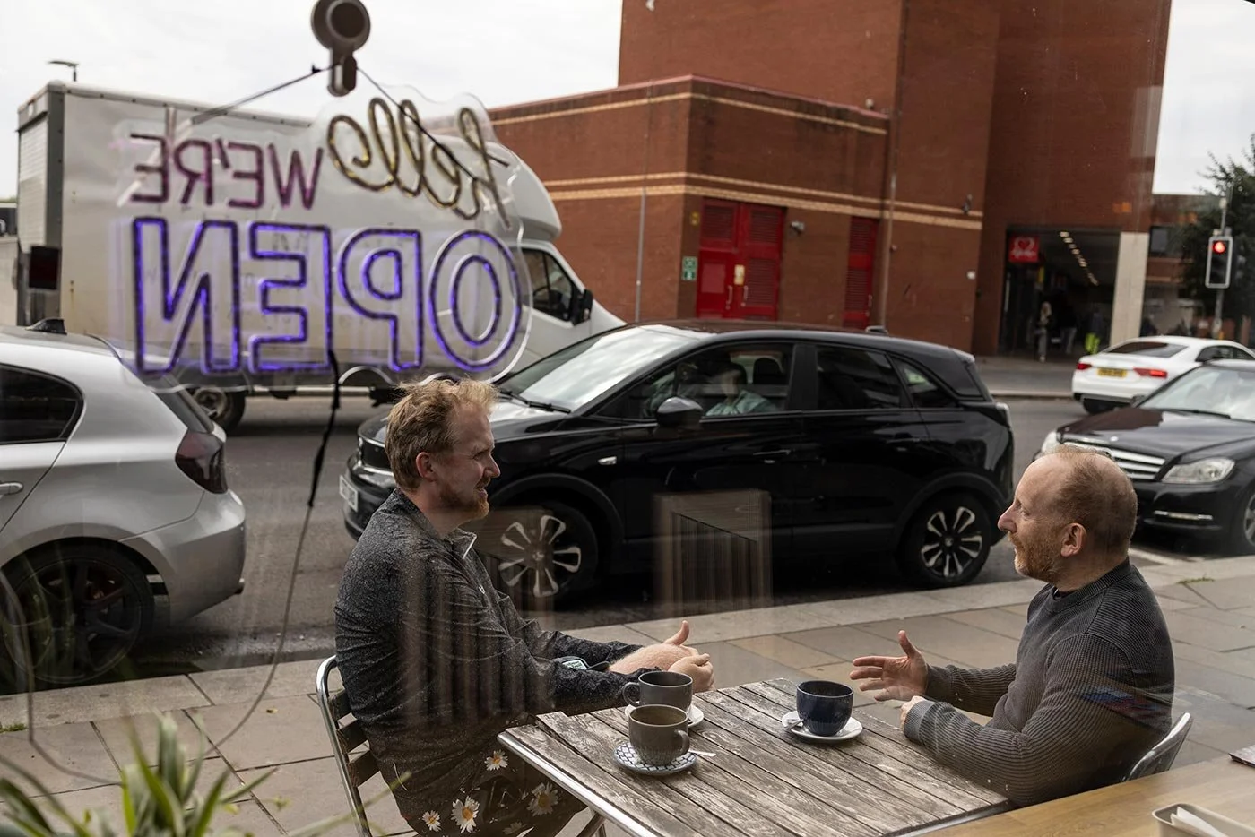 Two men sit outisde a coffee shop in conversation, they have coffee cups on the table before them. A neon sign reads (back to front in the window) We Are Open.