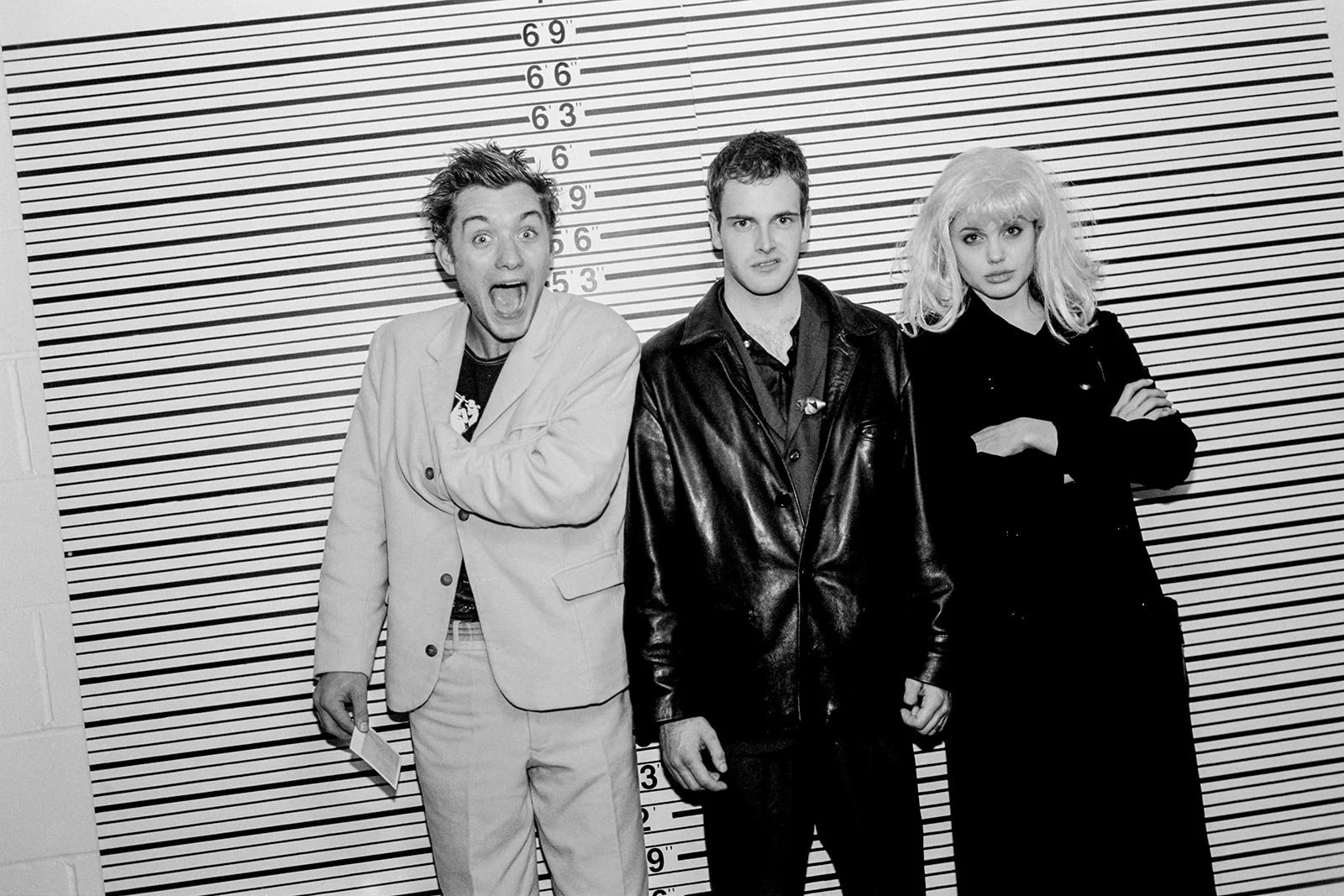 Two men, one in light suit making a funny face, and one in dark suit scowling stand in front of police line up wall, with woman wearing a blond wig. and a