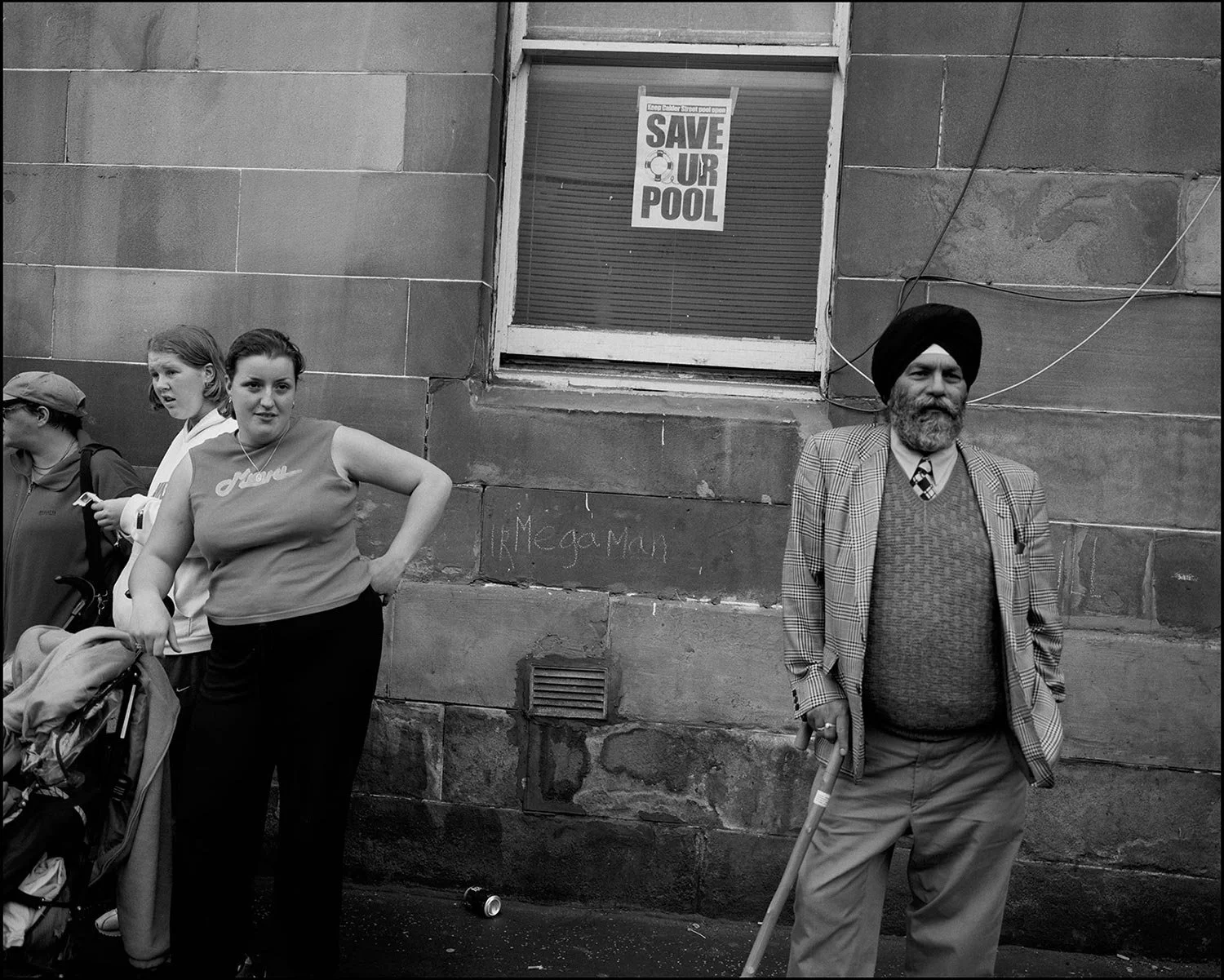 A young lady leans on a pram, beside to her right a Sikh man leaning on a walking stick, behind them on a window a poster reads Save OUr Pool. The photo is in black and white.