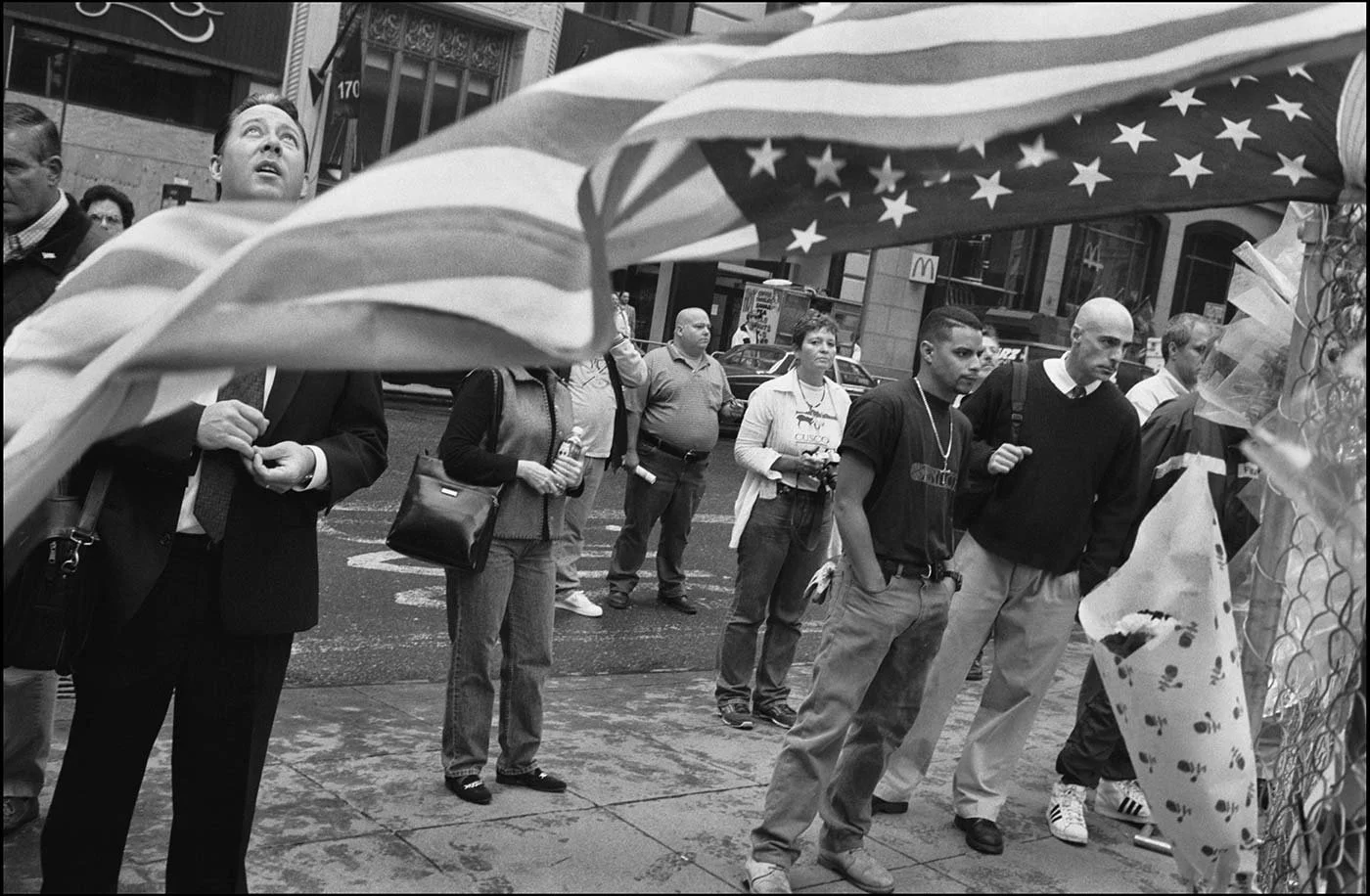 Black and white photo of people standing in a Manhattan street, all facing the same direction. An American stars and stripes flag blows across the image,