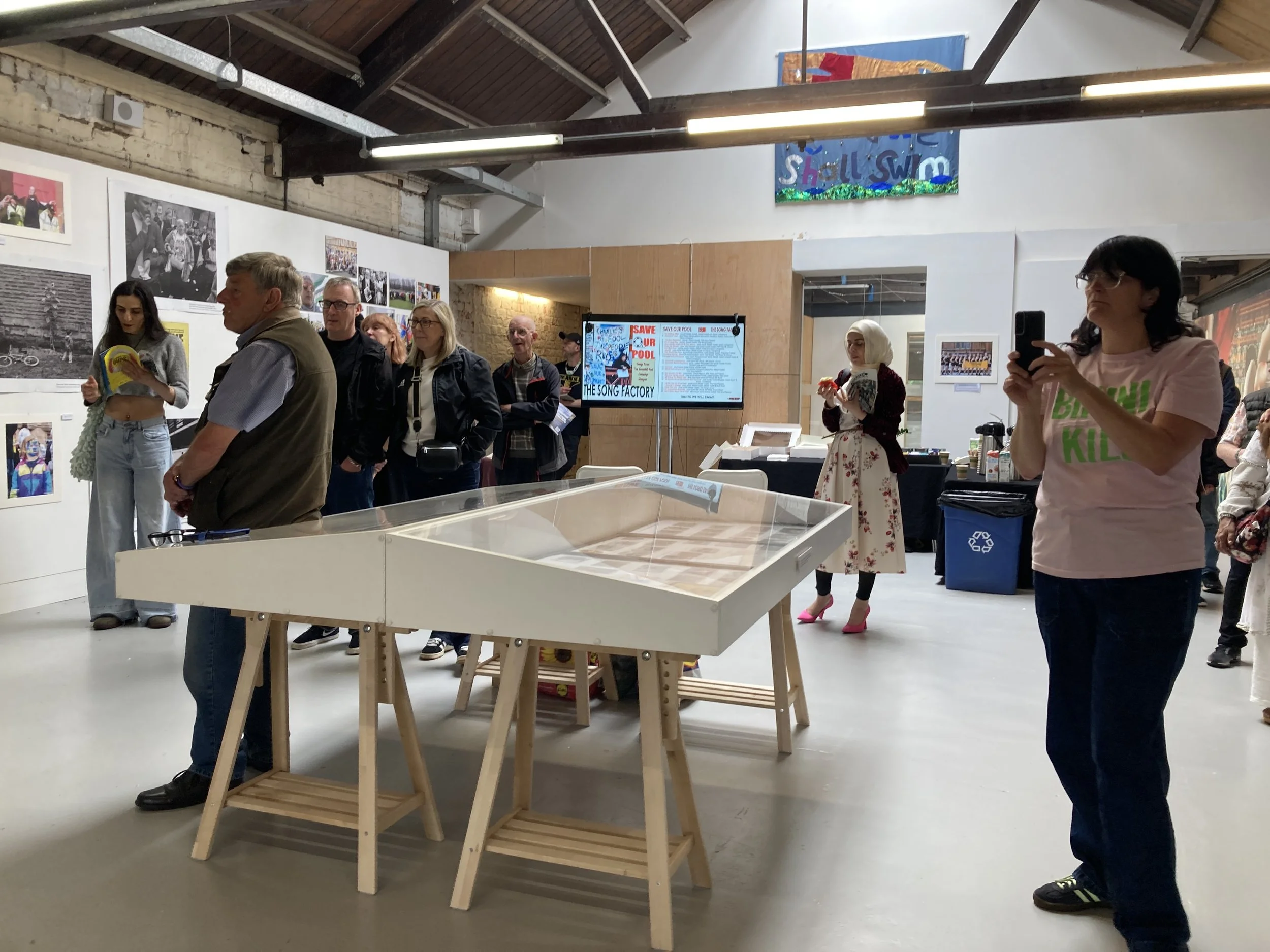 Colour photograph showing people to right of image socialisng at a photography exhibition. Black and white large photos are on the wall.