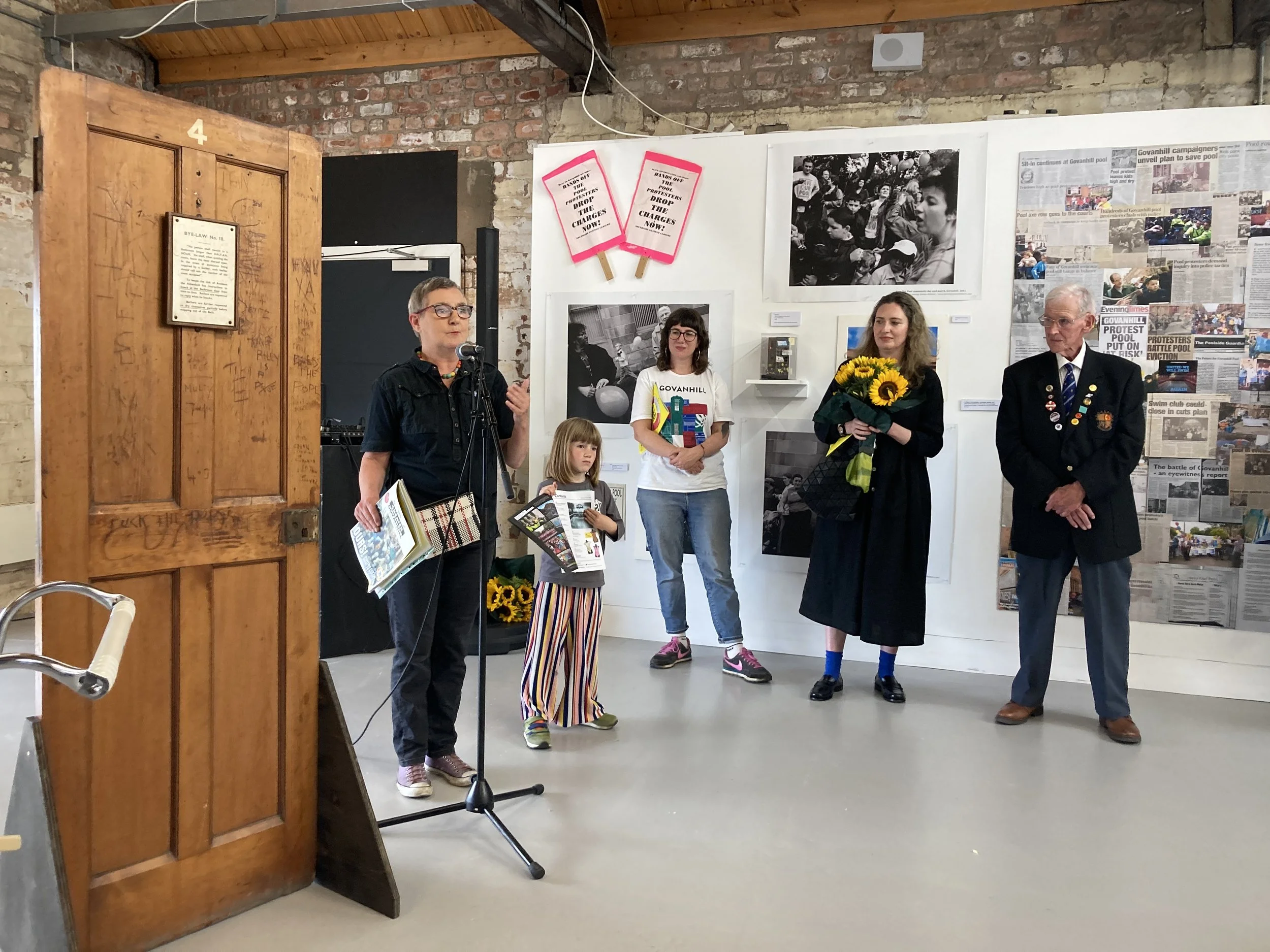 People stand in an exhibition, beside a wooden door on left of image, making speeches. There are four adults and one child.