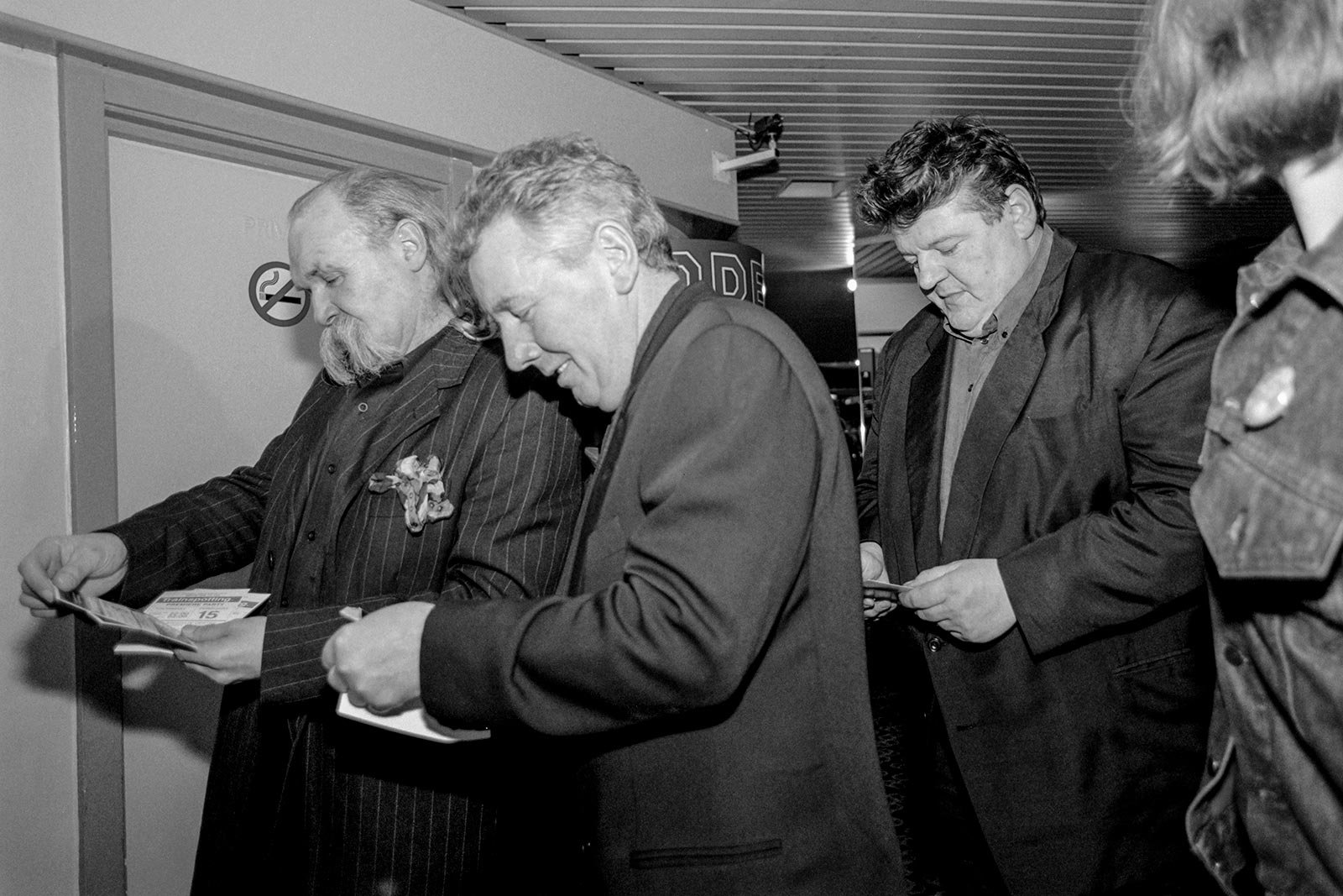 Three white middle aged men look at cinema tickets as they enter a room. They're all wearing suits and smiling.