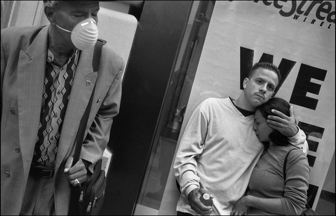 Black and white photo of a young man holding his grilfriend who is crying into his chest. To the left of picture a man in suit wears a face mask.