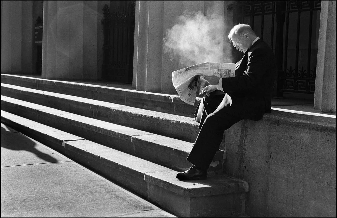 Black and white photo of a businessman sitting on stone steps, in front of columns, he smokes a cigar and reads a newspaper.