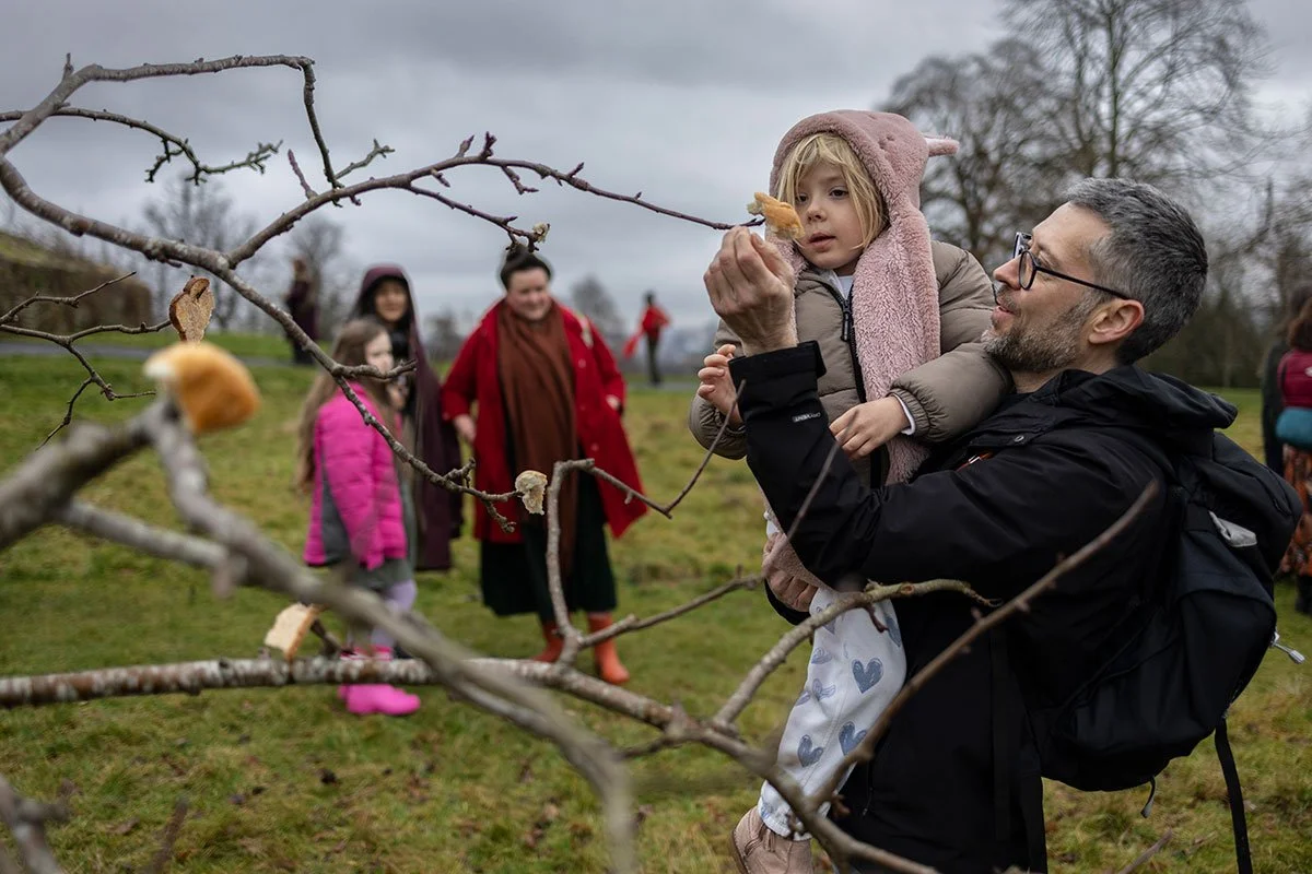A man holds a child and together they Affixing cider soaked bread to an apple tree, as way of an offering.