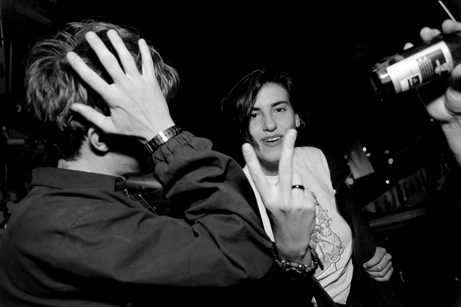 A man on the left of image covers his face with his hand, and a woman central in the image flashes a rude V sign at the camera as sshe confronts the photographer. The image is ina black and white and taken in a dark party using camera flash