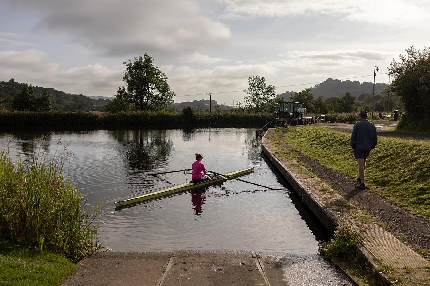 A man walks on a canal path, looking to his left at a woman on a rowing skul canoe on the water.