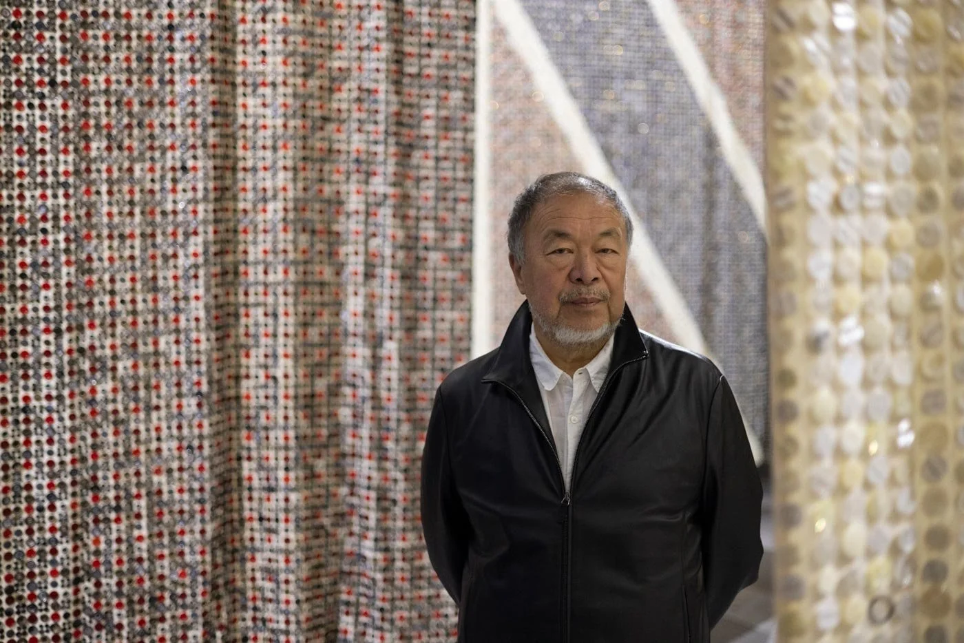 Chinese artist Ai Weiwei wearing a black leather jacket looks straight to camera, surrounded by his artworks made of buttons.