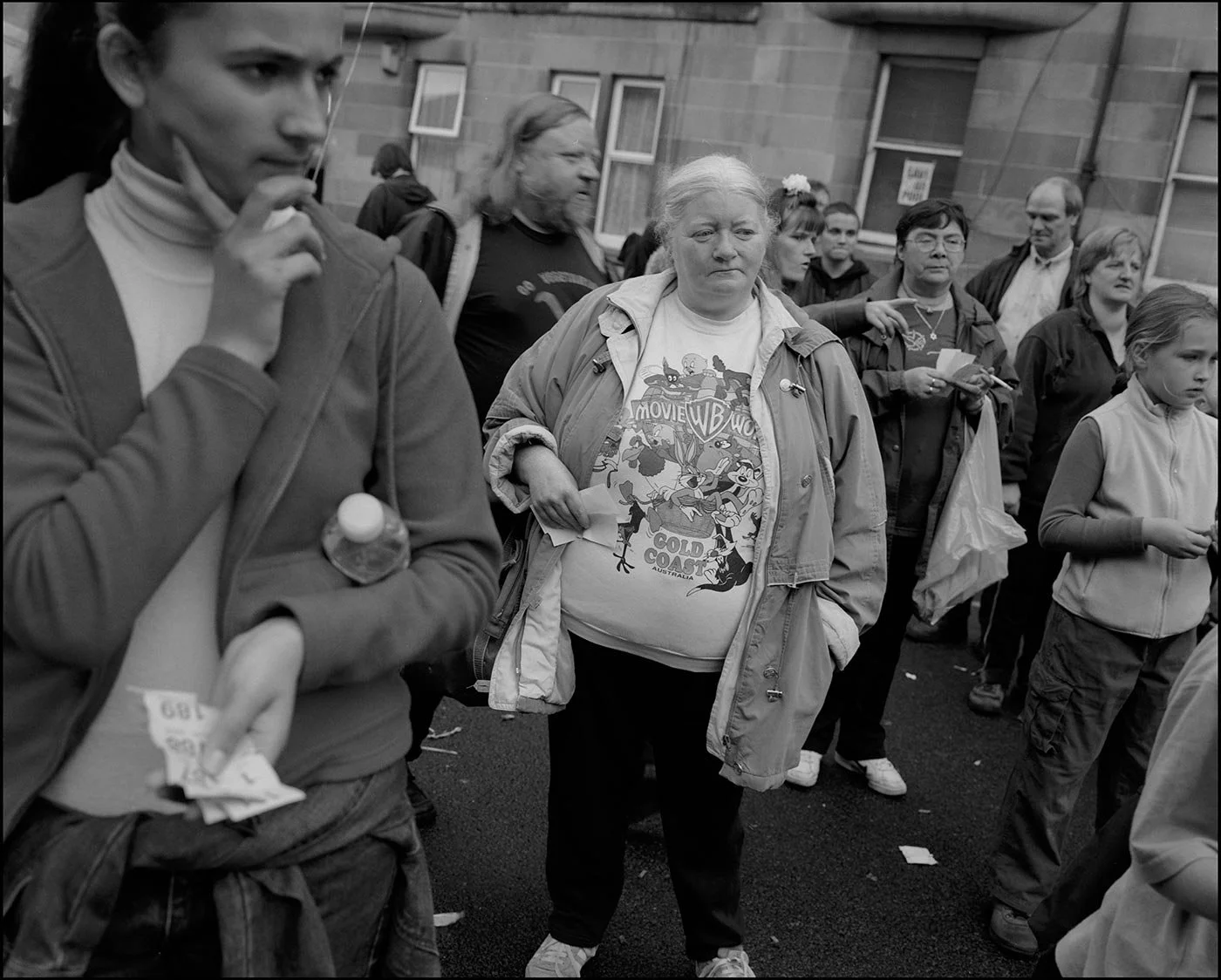 A black and white photo showing people standing in the street, facing camera, deep in thought. To the left a young Asian woman looks pensive. IN centre an older woman wearing a Warber Bros t-shirt, with cartoon characters on it.