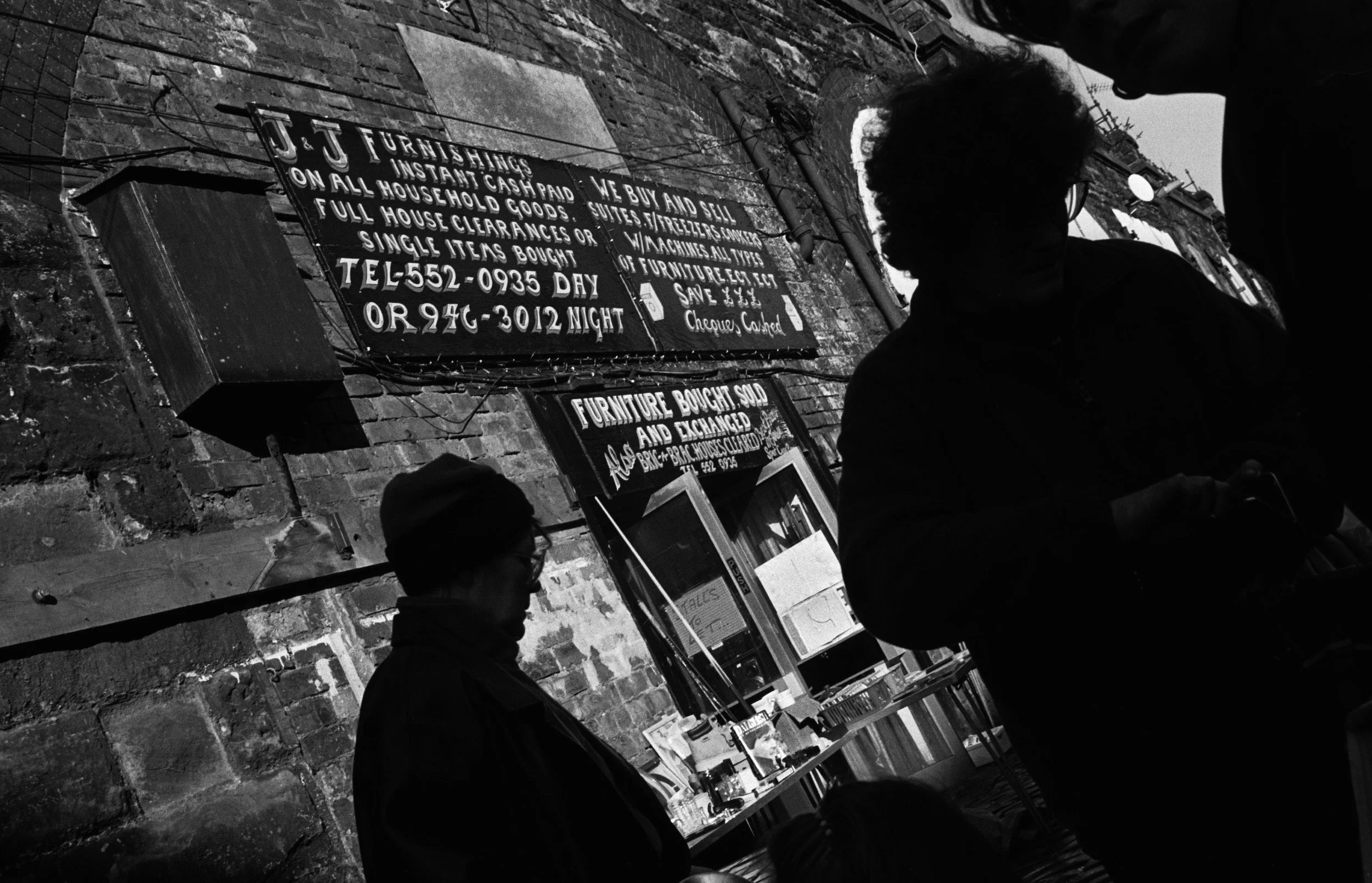 Silhouette of people in front of train railways arches, with signs on them from a flea market stall. The photogrpah sis horizontal in high contrast black and white.