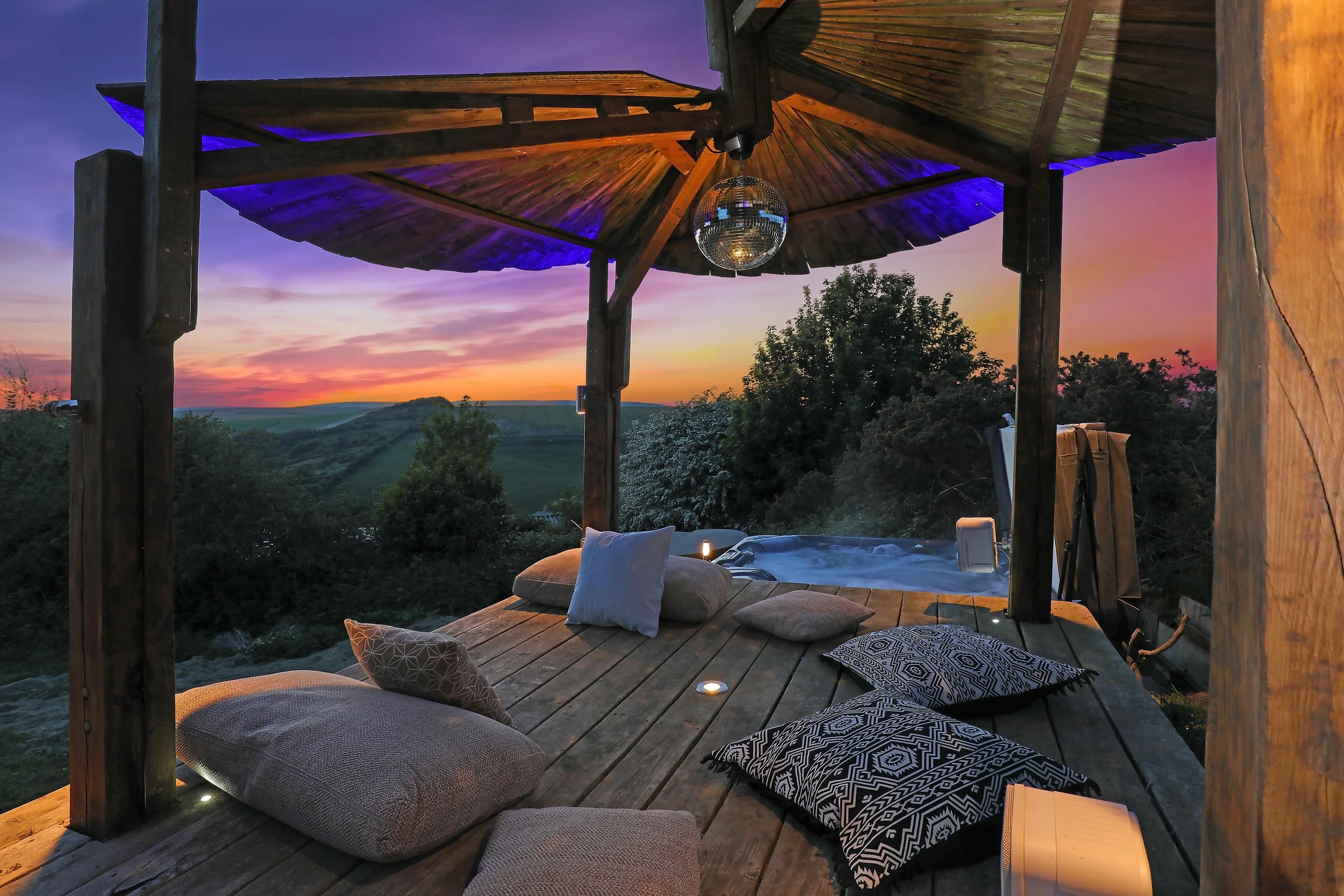 A cozy hot tub next to a rustic deck adorned with cushions and ambient lighting, offering breathtaking sunset views over rolling hills, perfect for relaxation during creative retreats.