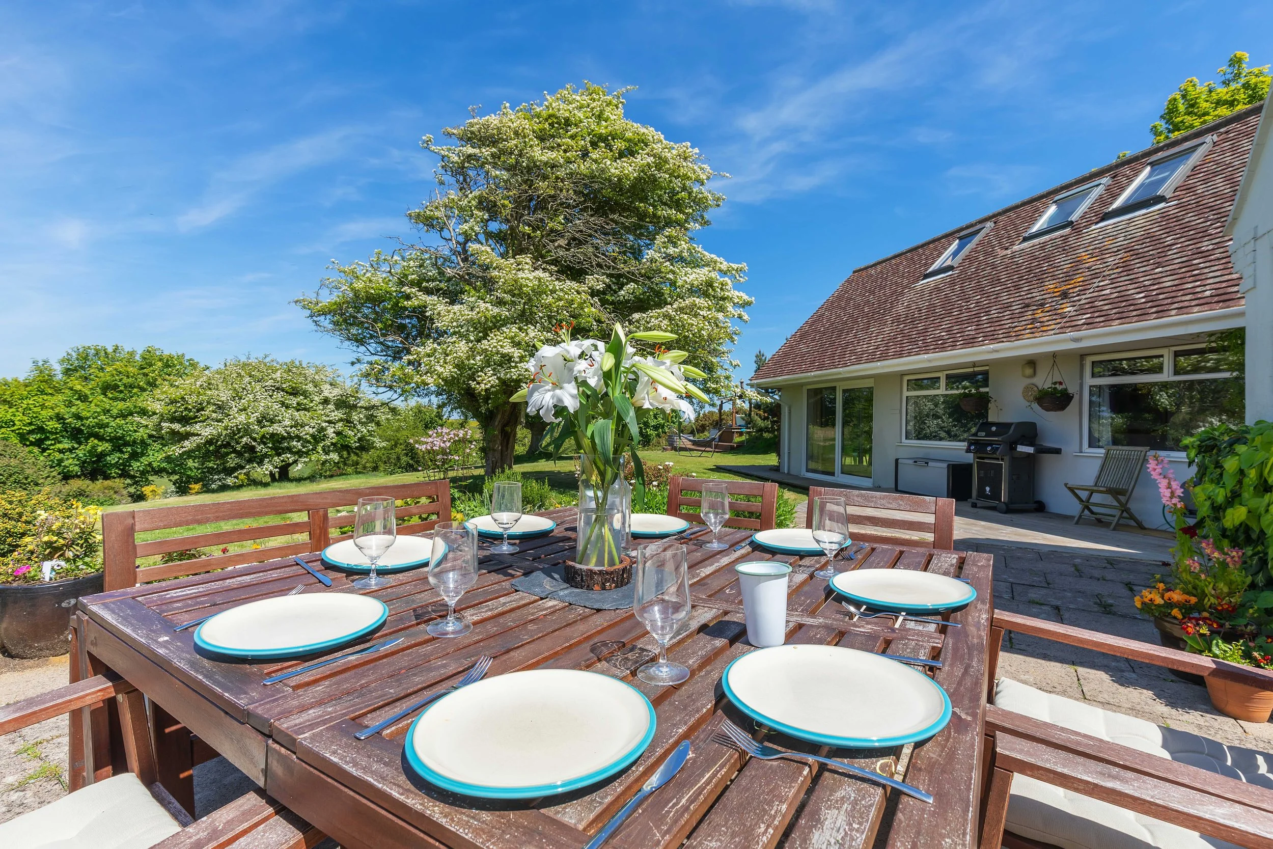 A beautifully set wooden dining table in a sunny outdoor garden, surrounded by lush greenery and flowering trees, with a charming retreat house in the background. Perfect for creative retreat outdoor gatherings and shared meals in a peaceful environm