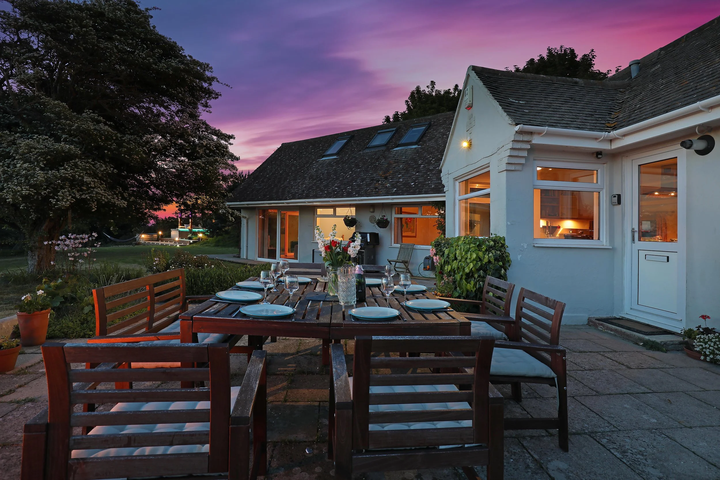 Outdoor dining area at a UK retreat accommodation, featuring a wooden table set for dinner under a stunning pink and purple sunset sky, surrounded by lush greenery and a cozy retreat cottage