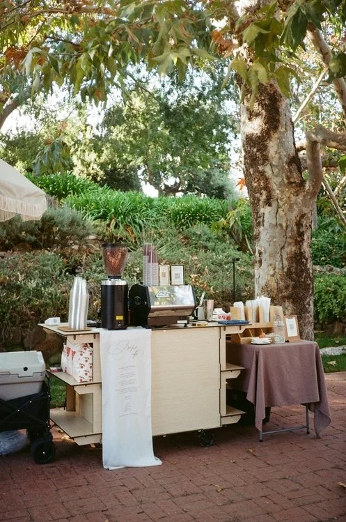 An outdoor wedding coffee cart stand with a La Marzocco GS3 espresso machine, cups, and supplies, set up under trees in a garden in Simi Valley, CA at Quail Ranch.