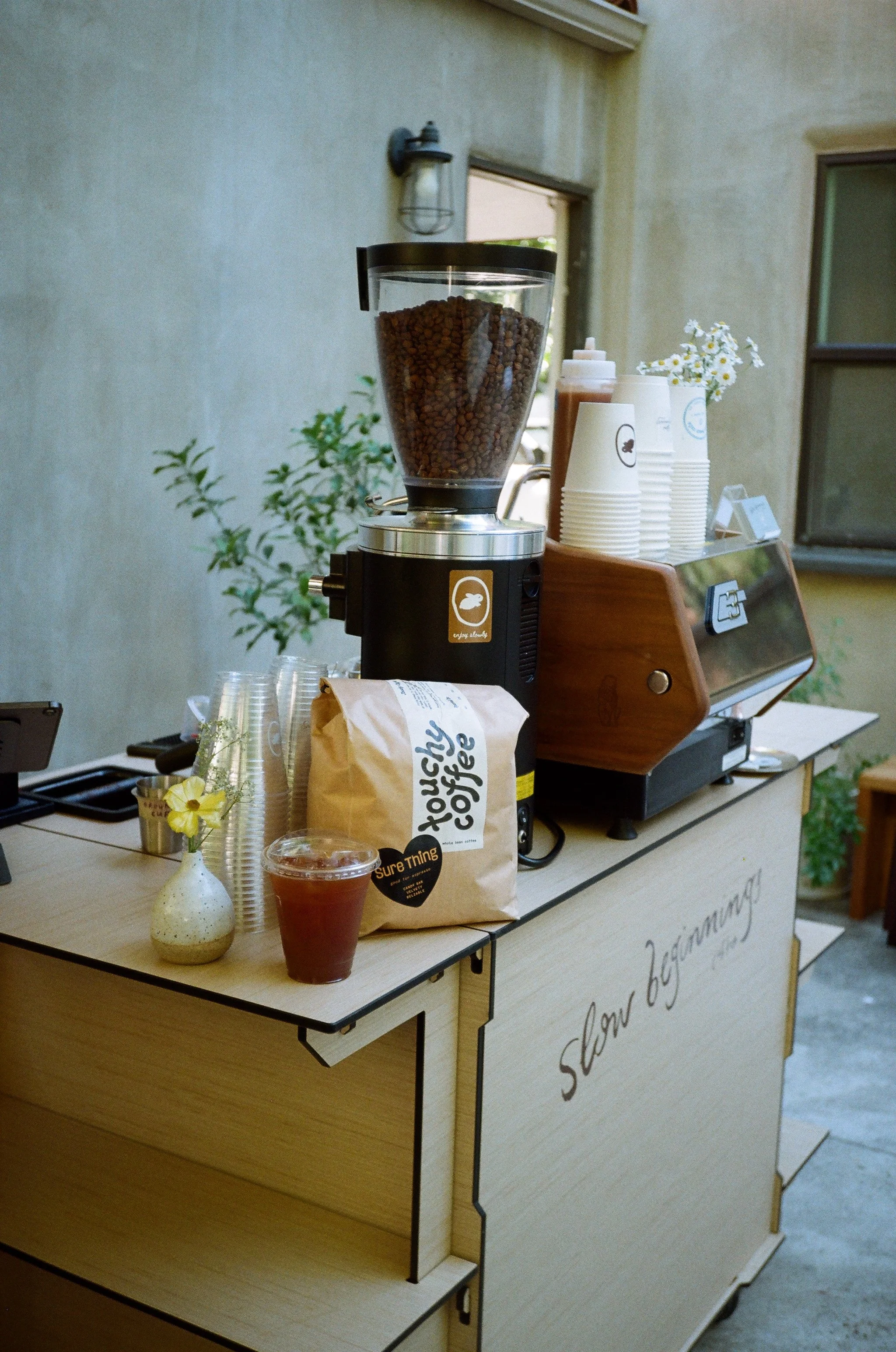 An outdoor wedding coffee cart stand with a La Marzocco GS3 espresso machine, cups, and supplies, set up under trees in a garden in Simi Valley, CA at Quail Ranch.