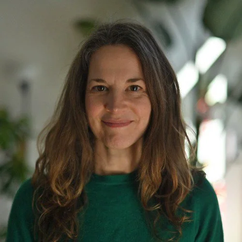 A woman with long, wavy brown hair wearing a green top, standing indoors with a blurred background of plants and windows.