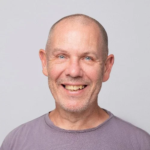 Smiling bald man with blue eyes and light facial hair, wearing a gray T-shirt, against a plain light background.