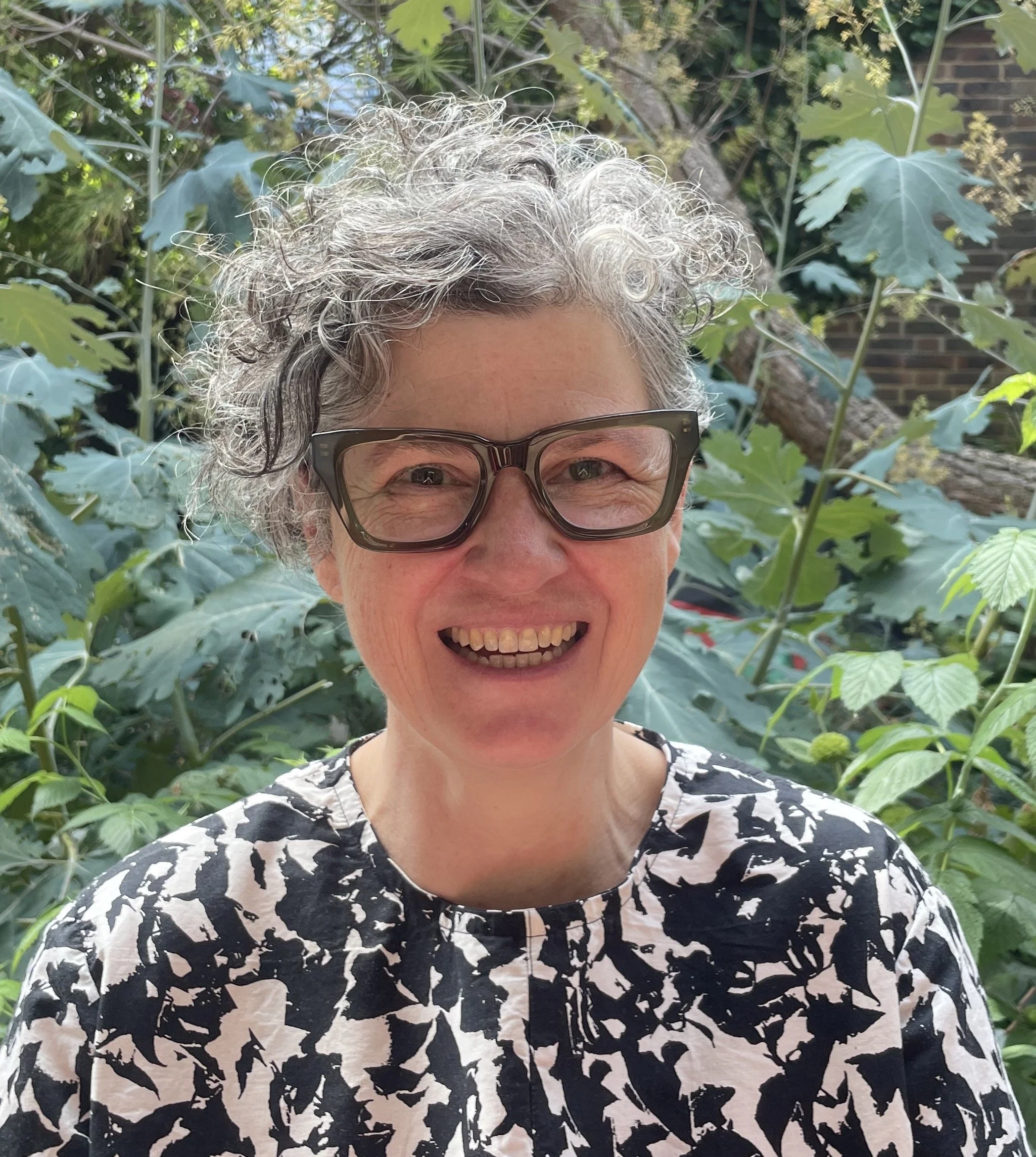 A smiling woman with curly gray hair and glasses, wearing a black and white patterned top, standing outdoors in front of lush green plants.