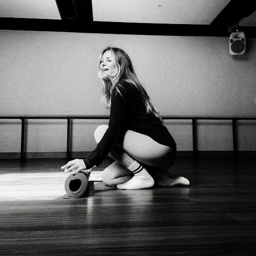 A black and white image of a woman in a long sleeved leotard and socks with a matt in a ballet studio 