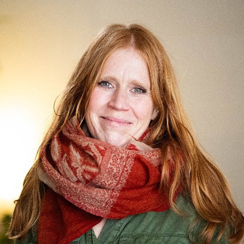 A woman with long red hair, wearing a red patterned scarf and green jacket, smiling at the camera.