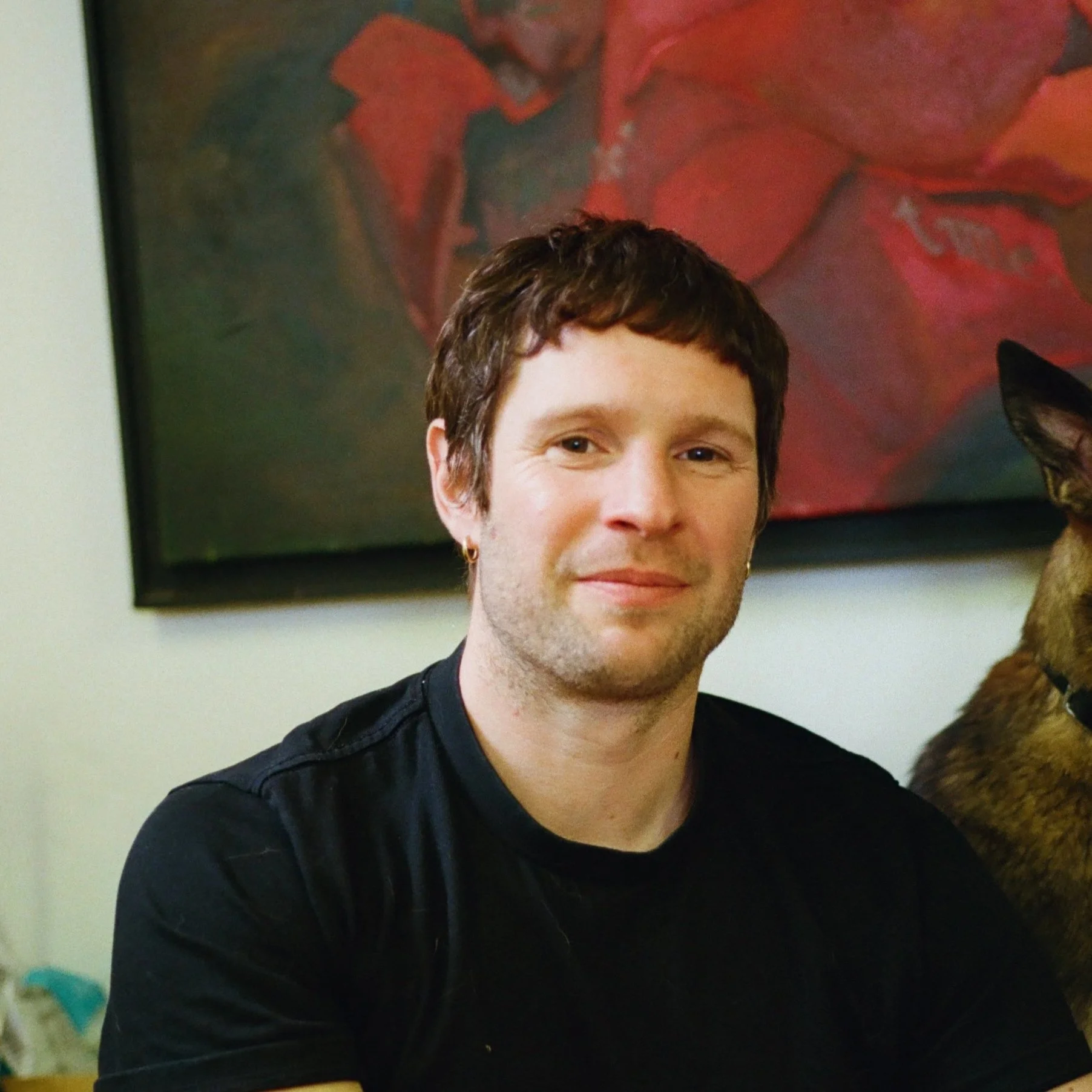 A person with short brown hair and a slight beard smiling at the camera, wearing a black shirt, sitting in front of a colorful abstract painting with a dog partially visible to the right.