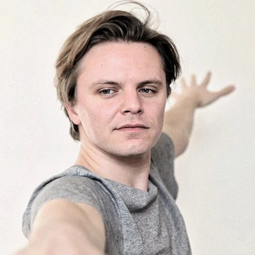 Man with light brown hair taking a selfie with a neutral expression, extending his arm toward the camera, against a plain light-colored wall.