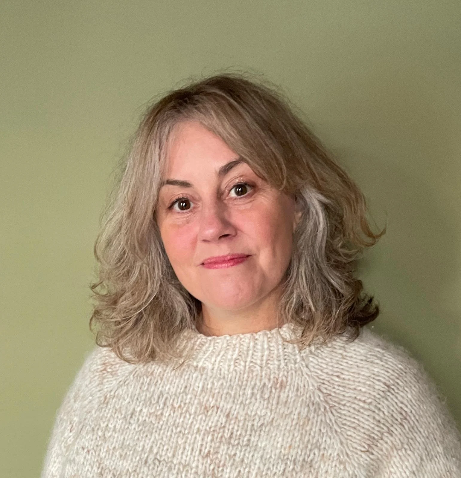 A woman with wavy, shoulder-length hair and light skin, wearing a white knit sweater, standing against a light green background.