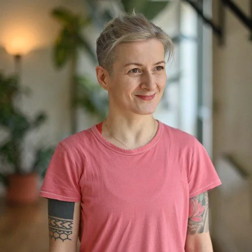 Woman with short gray hair, wearing a pink t-shirt, standing indoors with a smile, tattoos visible on both arms, background with plants and a staircase.