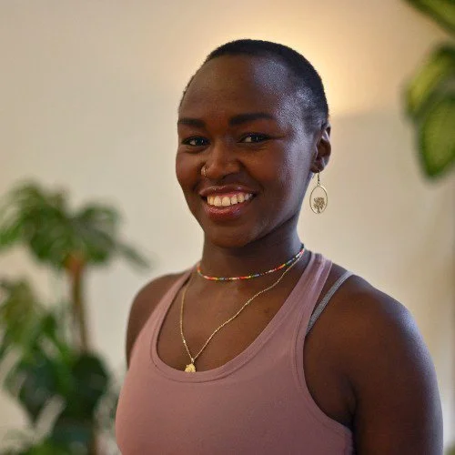 A smiling Black woman with short hair, wearing a pink tank top, colorful beaded necklace, and silver earrings with a tree design, standing indoors with green plants in the background.