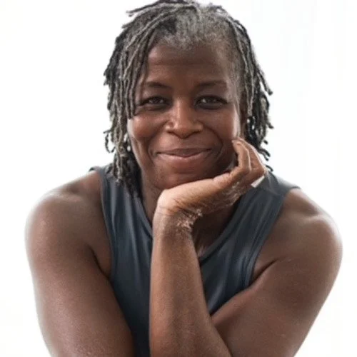 A woman with gray dreadlocks smiling and resting her chin on her hand, wearing a sleeveless gray shirt.